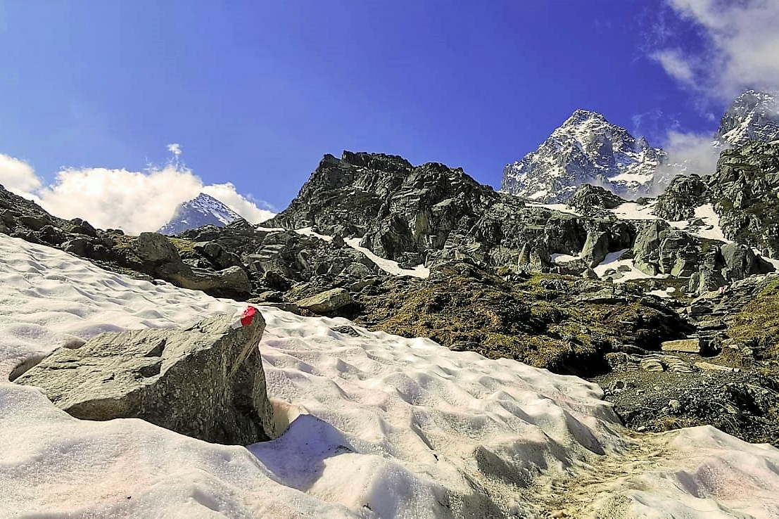 foto panoramica di montagna, in primo piano neve con una roccia con il segno bianco e rosso che emerge dalla neve, in secondo piano la catena montuosa dove si riconosce il monviso. cielo azzurro con qualche nuvola
