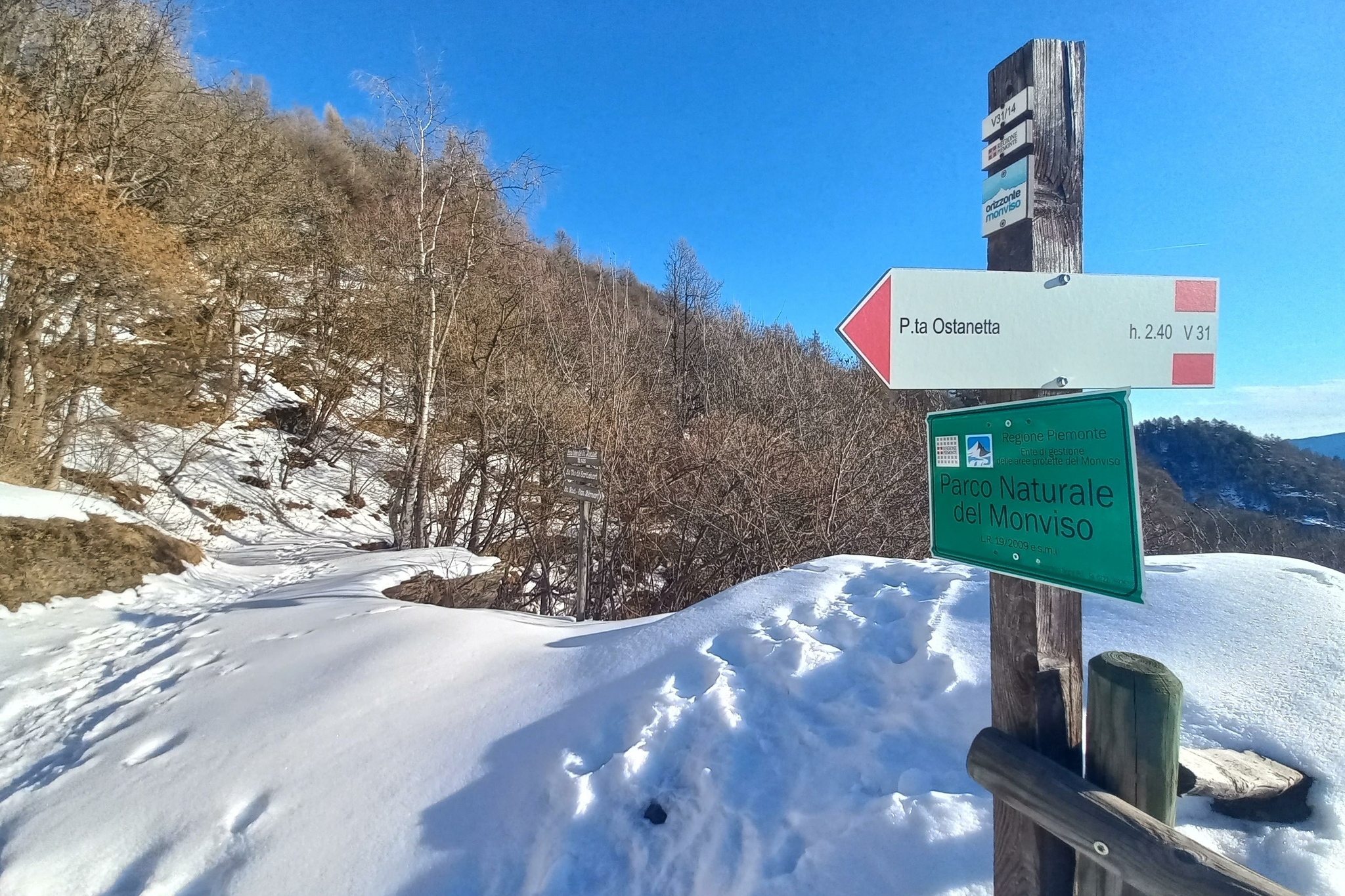 Palo con cartello verde indicante il confine del Parco naturale del Monviso e cartello segnavia bianco e rosso con la scritta P.ta Ostanette. intorno il sentiero innevato tra alberi in tenuta invernale, senza foglie. cielo azzurro
