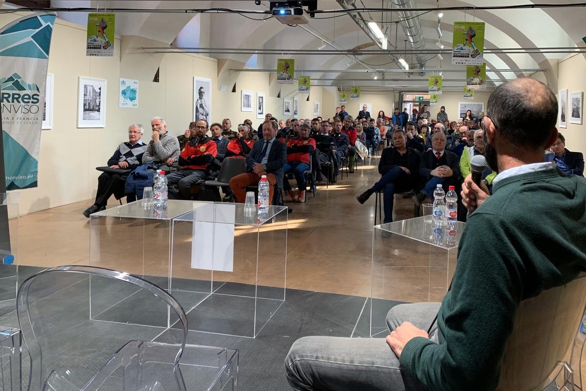 una persona di spalle con un microfono in mano, messa a destra nell'immagine, sta parlando alla sala piena di persone.