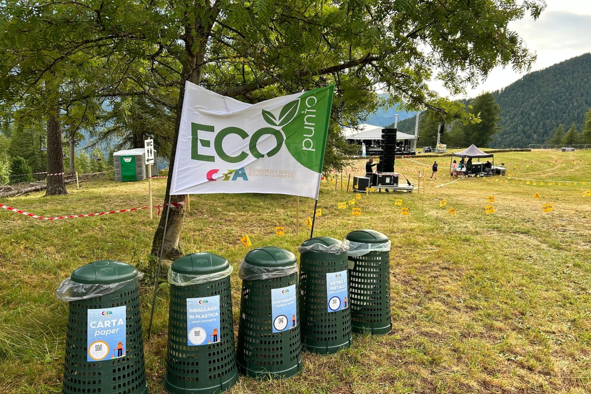 Cinque contenitori per rifiuti differenziati in plastica verde in un prato; sullo sfondo si vede il palco di un evento musicale in corso di organizzazione (foto di repertorio)