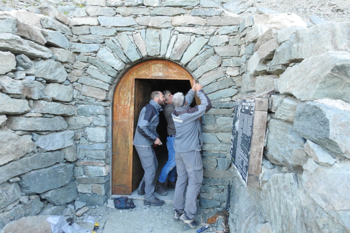 artigiani al lavoro per posizionare la nuova porta al Buco di Viso: tre persone stanno fissando i cardini della porta con un trapano, si vede l'imbocco francese del tunnel.