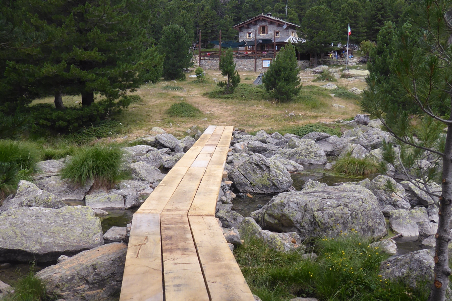 In un bosco, in basso al centro della foto si vede la nuova passerella installata nei pressi del lago Bagnour: in lontananza si scorge l'edificio del rifugio omonimo.