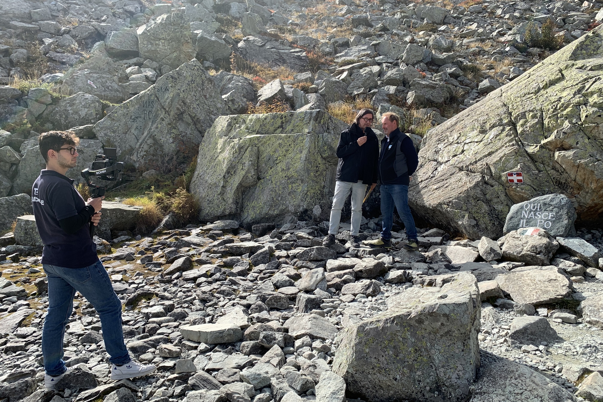 Alle sorgenti del Po, visibili sulla destra dell'immagine, un cameraman sta inquadrando due persone che parlano: il conduttore Claudio Calorio e il presidente del Parco del Monviso Dario Miretti.