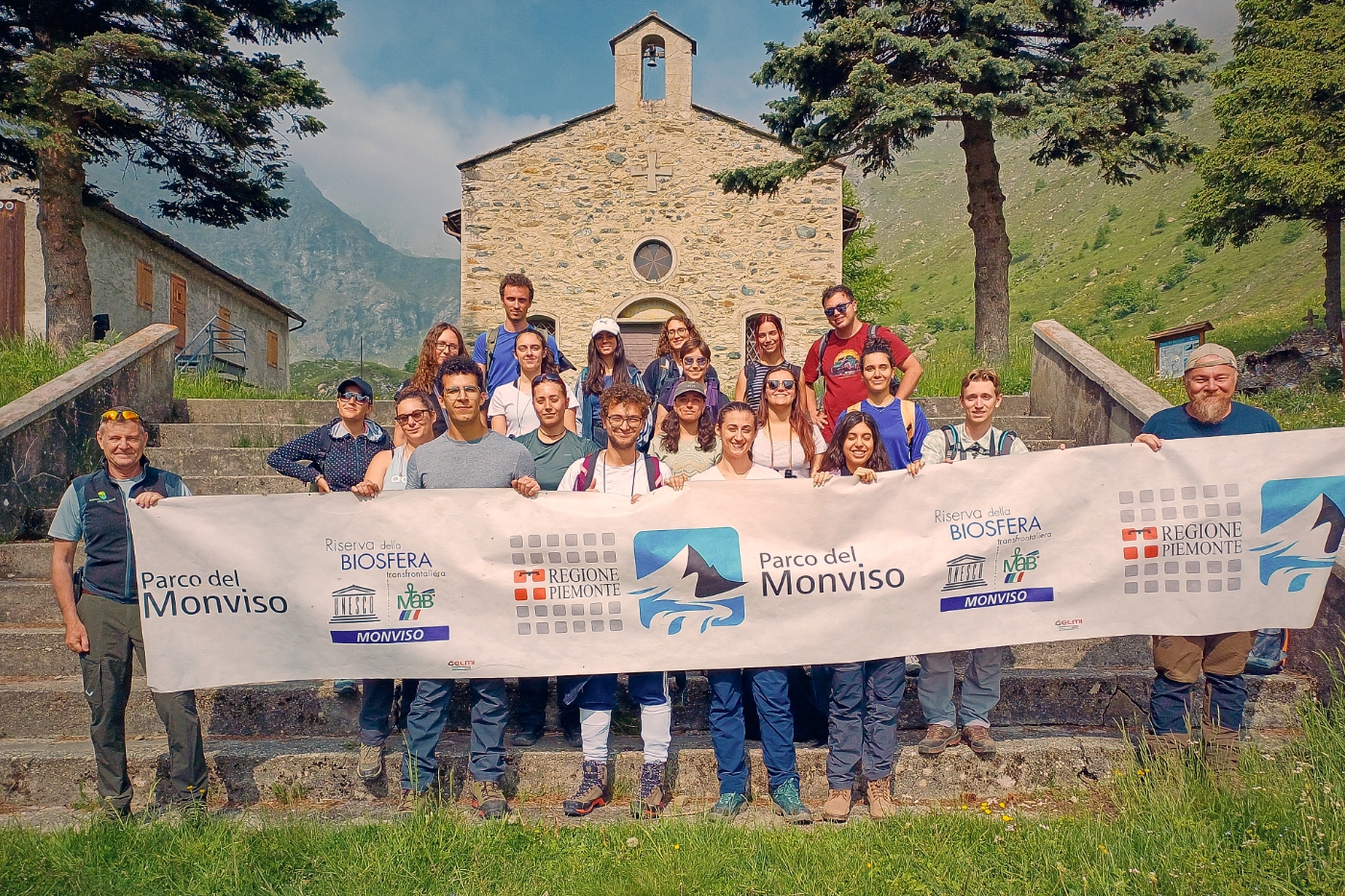 foto di gruppo in contesto montano: il gruppo di persone tiene uno striscione con il logo del Parco del Monviso davanti a una chiesetta montana