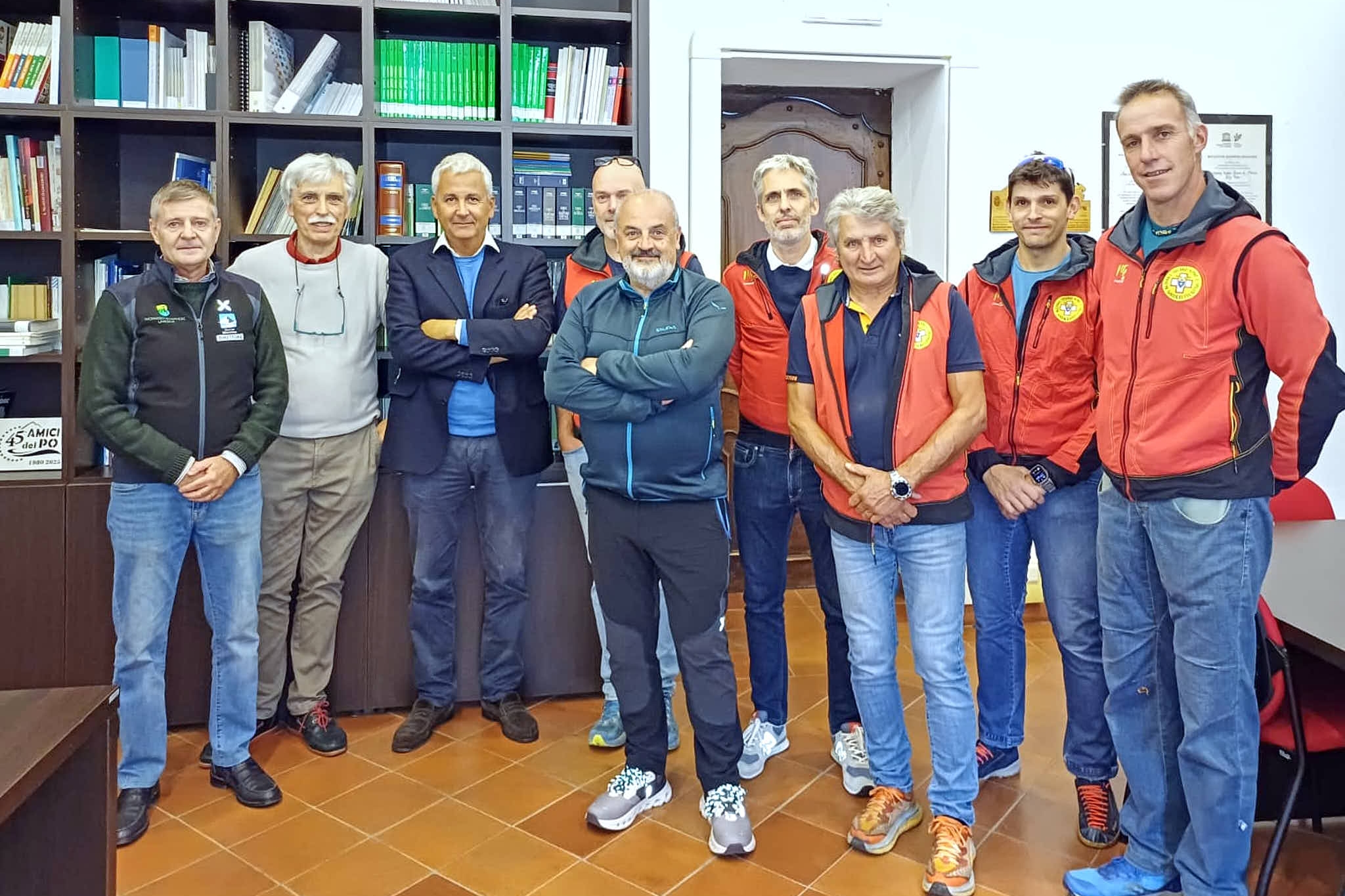 Foto di gruppo di nove uomini in piedi in un ufficio della sede di Saluzzo del Parco del Monviso. Il gruppo è composto da persone di età variabile, prevalentemente di mezza età. Quattro di loro indossano giacche rosse e blu del Soccorso Alpino, riconoscibili dal logo circolare giallo sul petto. Gli altri indossano abbigliamento casual o sportivo: giacche blu, felpe e jeans. Tutti sorridono guardando verso la fotocamera in un'atmosfera cordiale e informale. Sullo sfondo si vede una libreria a scaffali scuri piena di volumi rilegati di vari colori, tra cui spiccano alcuni dorsi verdi allineati. Sulla destra è visibile una porta bianca con un vecchio mobile scuro all'interno e alcune targhe e certificati appesi alla parete. Il pavimento è in piastrelle color terracotta.