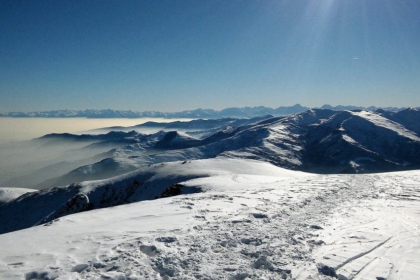 Dalla cima di una montagna panorama delle montagne circostanti completamente innevate al di sopra di una distesa di nuvole sulla sinistra. Cielo azzurro