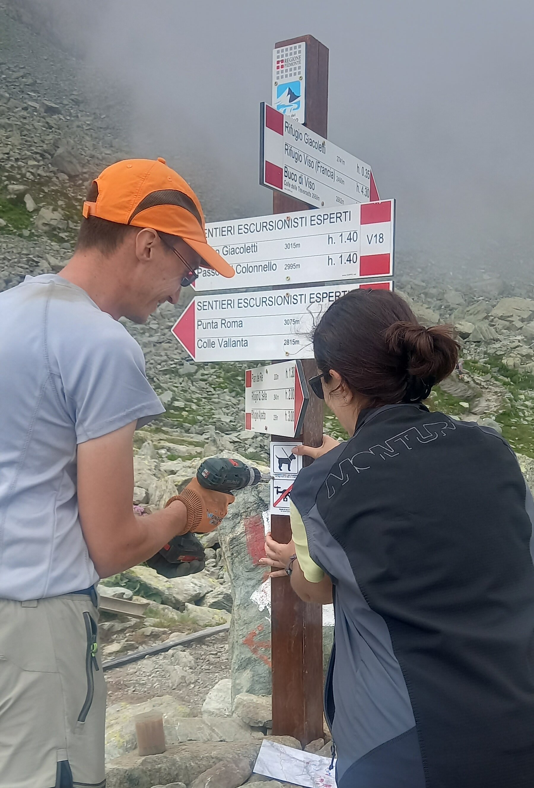 Due operatori del Parco del Monviso sono impegnati nella manutenzione della segnaletica escursionistica in alta montagna. Una persona utilizza un avvitatore per fissare un cartello direzionale, mentre l'altra lo mantiene in posizione. Il palo riporta numerose indicazioni per rifugi, passi e itinerari escursionistici, tra cui il Rifugio Giacoletti, il Rifugio Viso, il Buco di Viso, Punta Roma e il Colle Vallanta. Sullo sfondo si estende un ambiente roccioso d'alta quota, con sentieri tra i detriti e nubi basse che avvolgono il paesaggio.