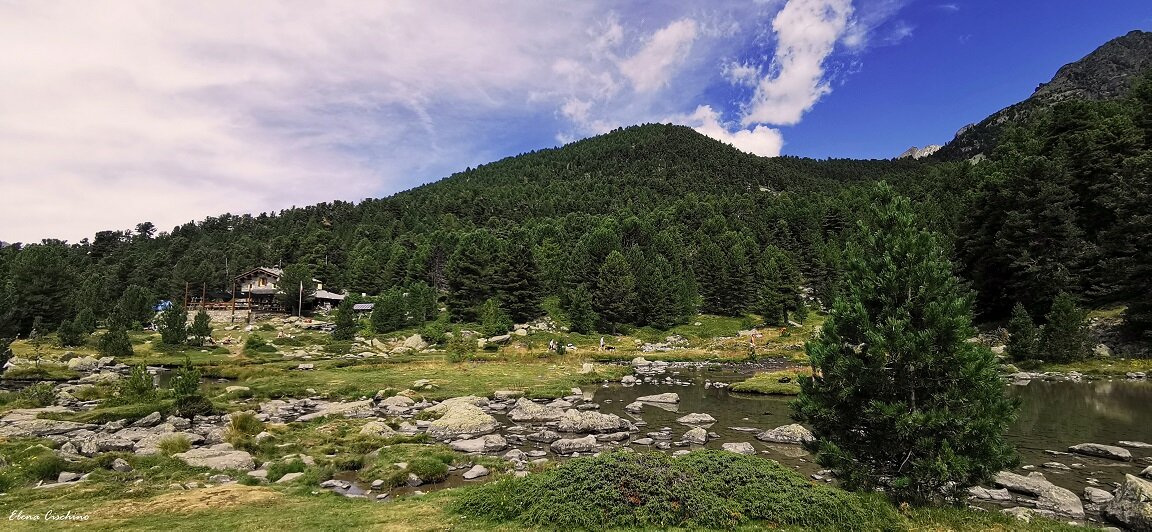 tra le rocce in primo piano si vede il lago bagnour, sulla sinistra il rifugio su un rialzo in pietra, in primo piano a destra un pino cembro. cielo azzurro con nuvole