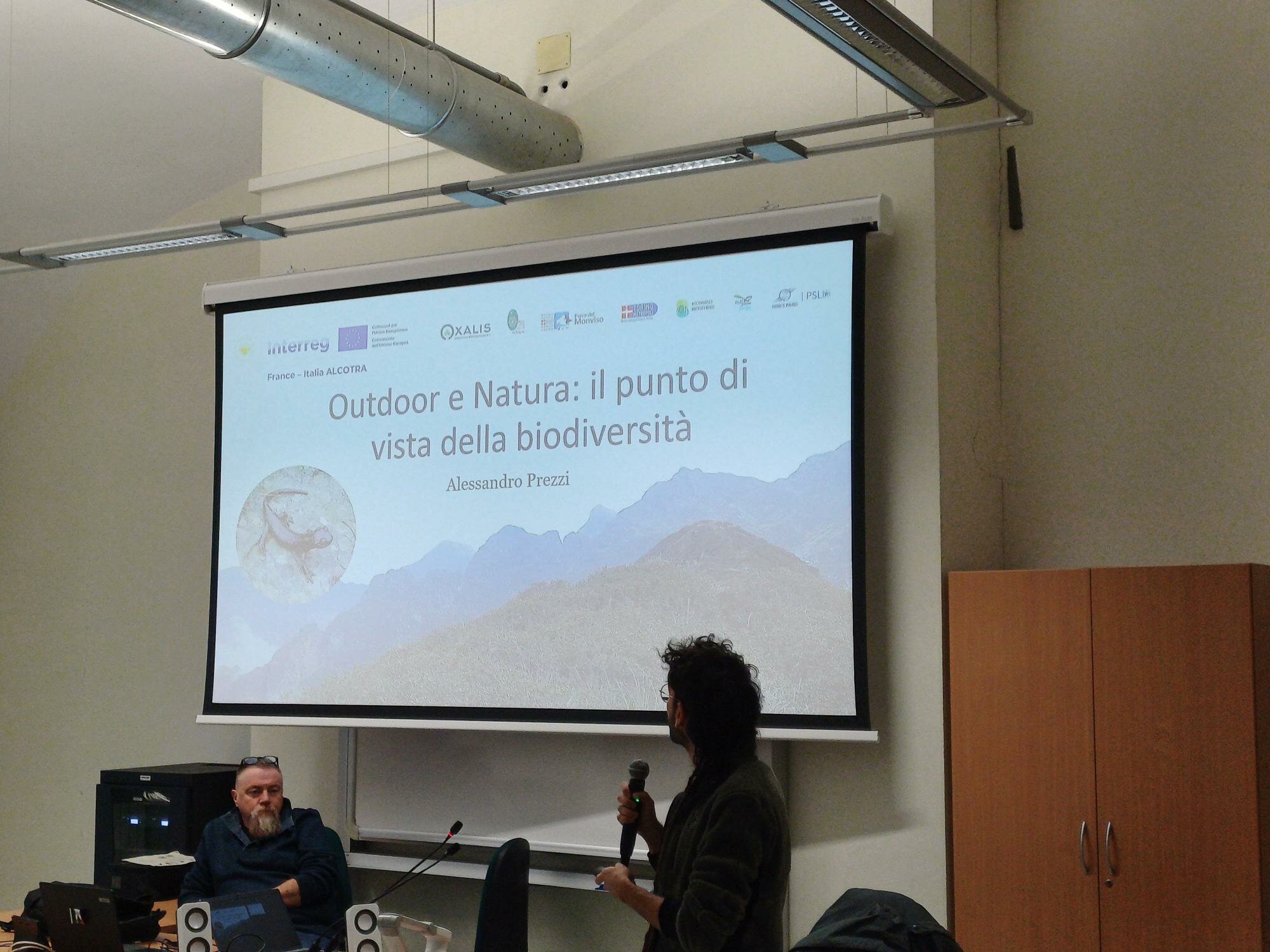 In un’aula universitaria, un tecnico del Parco del Monviso presenta al microfono davanti a uno schermo che mostra una diapositiva intitolata “Outdoor e Natura: il punto di vista della biodiversità” di Alessandro Prezzi. Seduto al tavolo, il professor Stefano Fenoglio osserva la scena. Sullo schermo compaiono loghi di vari enti e, sullo sfondo della slide, un’immagine montana. L’ambiente è semplice, con attrezzatura informatica sul tavolo e un armadio di legno sulla destra.