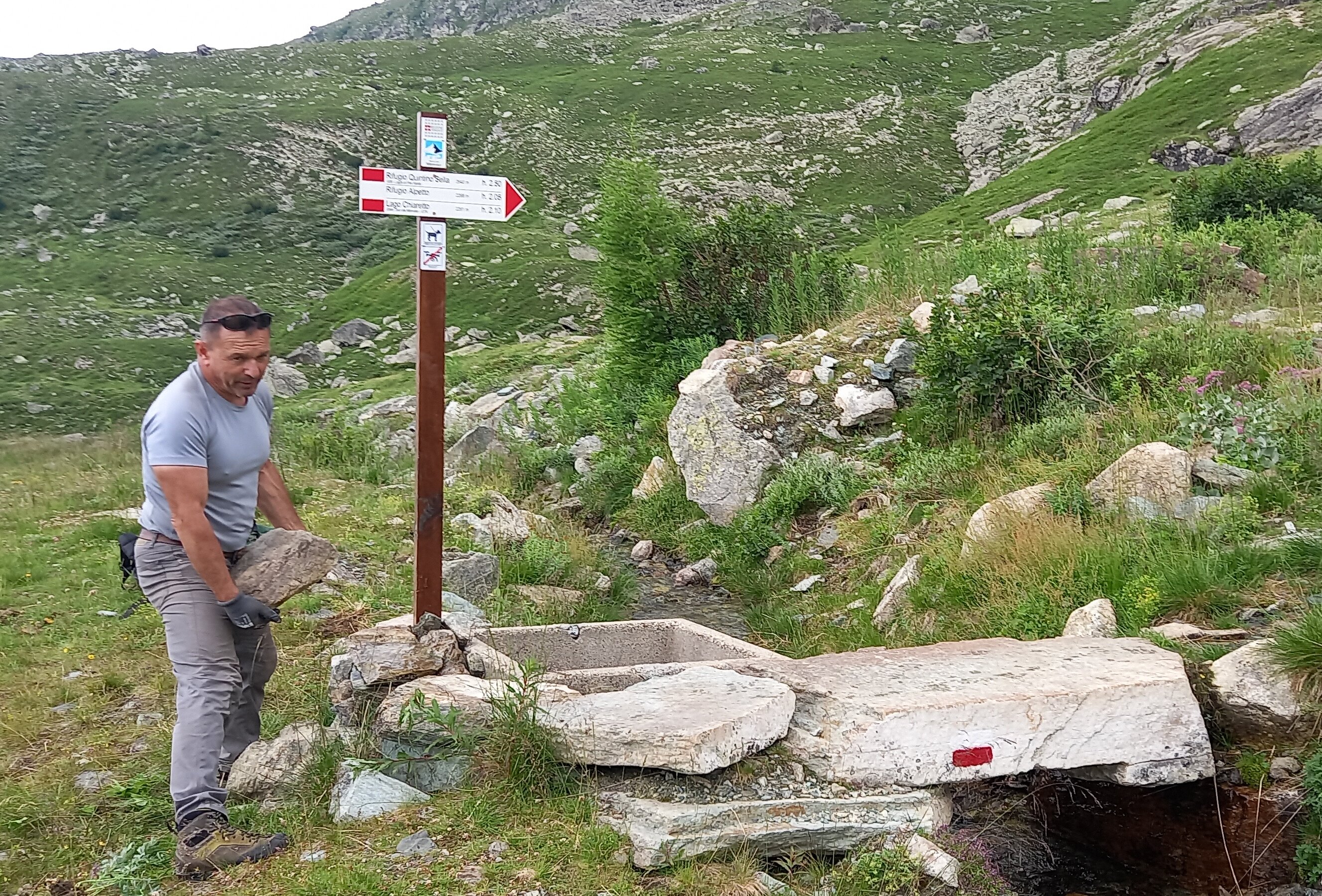 Un operatore del Parco del Monviso è colto mentre solleva una grossa pietra nei pressi di un palo della segnaletica escursionistica con indicazioni per il Rifugio Quintino Sella, il Rifugio Alpetto e il Lago Chiaretto. L'area è circondata da prati alpini, rocce e cespugli, con un piccolo corso d'acqua che scorre accanto.