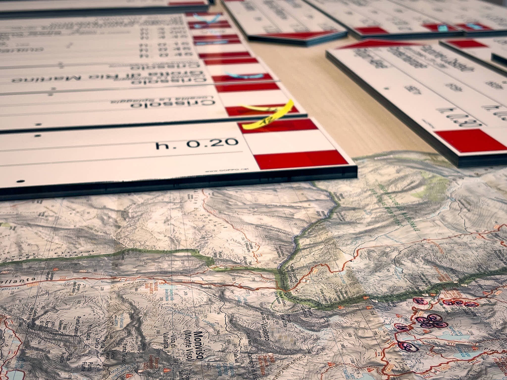 Dettaglio di materiali cartografici e informativi disposti su un tavolo da lavoro. In primo piano si vede una carta topografica dettagliata dell’area montana intorno al Monviso con curve di livello, sentieri tracciati in rosso e verde, toponimi e numerazione dei percorsi. Sopra la mappa sono appoggiate diverse schede o cartoncini rigidi di formato allungato con bordi rossi e informazioni stampate. Le schede sono disposte in modo ordinato, alcune sovrapposte ad altre.  L'insieme suggerisce un lavoro di pianificazione o catalogazione di percorsi escursionistici, probabilmente nell'ambito del progetto di valorizzazione del Parco del Monviso attraverso manutenzione e ripristino dei sistemi di tracciabilità e informazione per escursionisti.