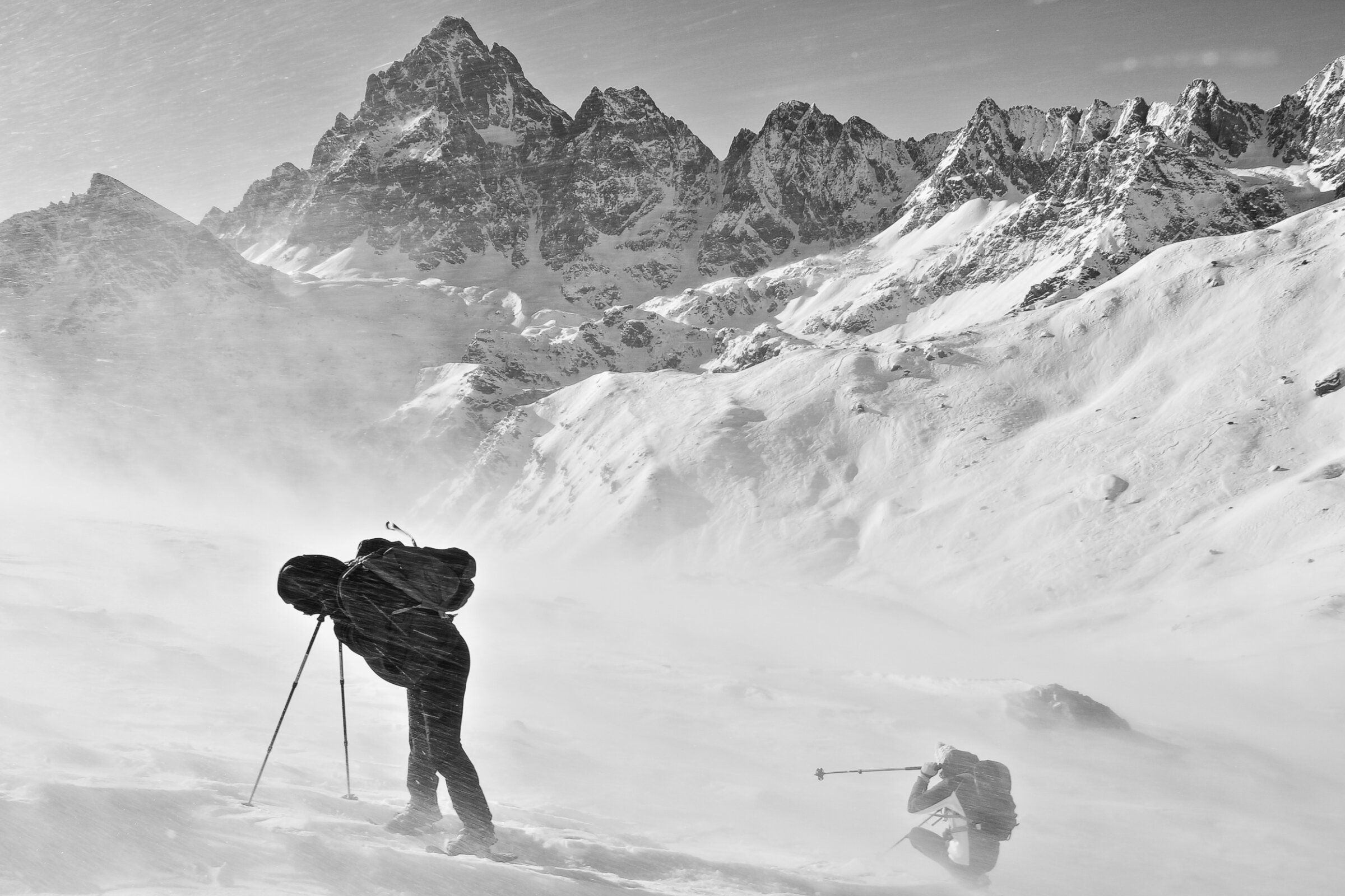 due scialpinisti investiti da un forte vento che solleva neve, sullo sfondo si vede la piramide del Monviso. Immagine in bianco e nero