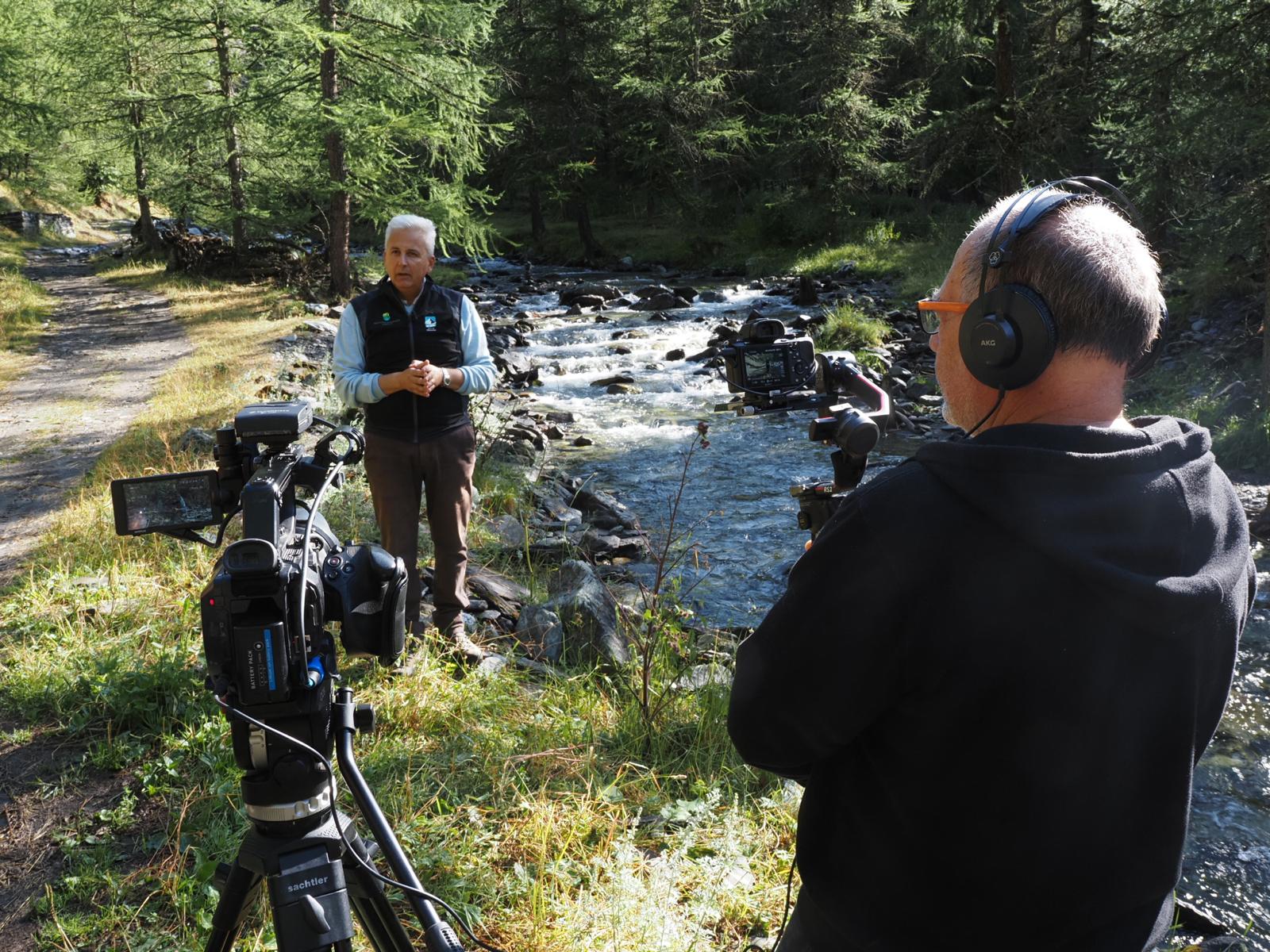 in primo piano un cameraman di schiena sulla destra della foto, sulla sinistra una seconda telecamera su treppiede; sullo sfondo un uomo di circa 60 anni che parla, inquadrato. Il contesto è naturale, a fianco delle persone scorre un torrente e ci sono alberi tutto intorno.