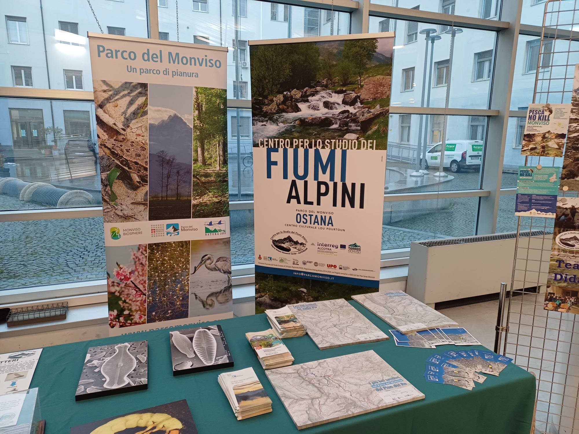 Un tavolo espositivo, allestito in un ambiente luminoso con grandi finestre, mostra materiale informativo del Parco del Monviso e del Centro per lo Studio dei Fiumi Alpini di Ostana. Sul fondo si vedono due roll-up: uno dedicato al Parco del Monviso, con immagini naturalistiche, e uno dedicato ai fiumi alpini, con la foto di un torrente di montagna. Sul tavolo sono disposti opuscoli, mappe, brochure e alcuni pannelli illustrati. Alla destra dell’esposizione, una griglia metallica sostiene altri poster informativi.