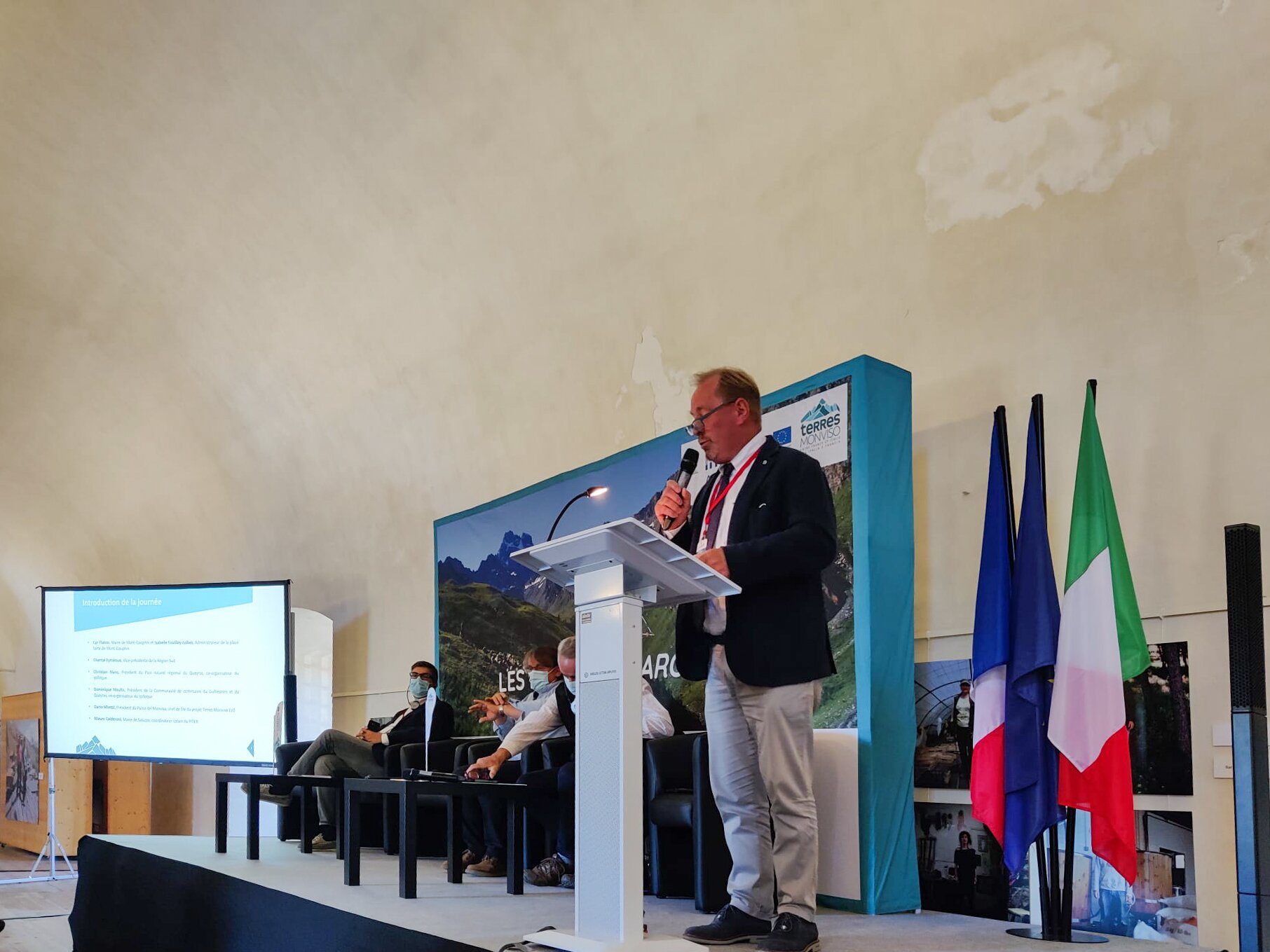 Il presidente del Parco del Monviso, Dario Miretti, durante la lettura del suo intervento: un uomo in giacca e cravatta davanti ad un leggio, a fianco delle bandiere italiana, francese e europea.