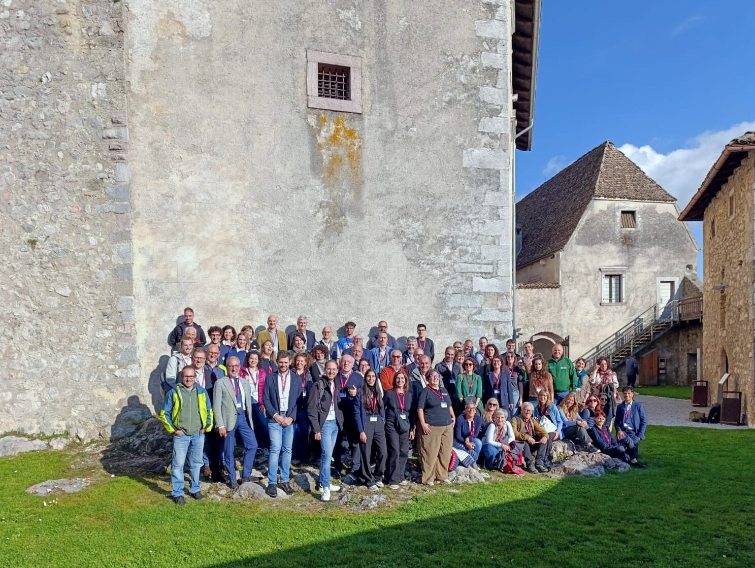 Nell’immagine un grande gruppo di persone posa per una foto di gruppo all’aperto, davanti a un edificio in pietra di aspetto storico. I partecipanti, circa un’ottantina, sono disposti su più file e molti indossano badge o cartellini identificativi al collo, segno di un incontro o convegno ufficiale. Il cielo è sereno e la luce del sole illumina la scena, creando un’atmosfera conviviale e ordinata in un contesto architettonico di pregio.