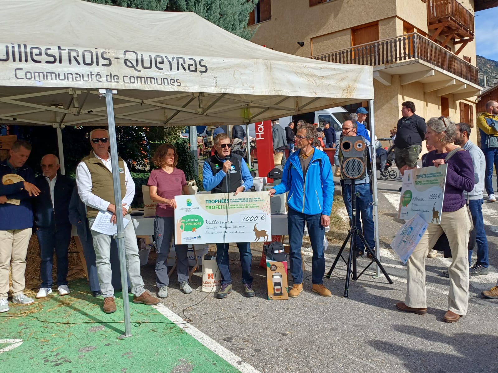 Cerimonia di premiazione dei Trofei della Riserva della Biosfera transfrontaliera del Monviso MaB UNESCO, organizzata all'aperto sotto un gazebo della Comunità di Comuni del Guillestrois-Queyras. Al centro, un uomo parla al microfono mentre due persone mostrano un grande assegno simbolico da mille euro. Sulla destra, una donna tiene un secondo assegno dello stesso importo. Altri partecipanti assistono alla cerimonia, sullo sfondo di edifici di montagna con balconi in legno, in una giornata soleggiata.