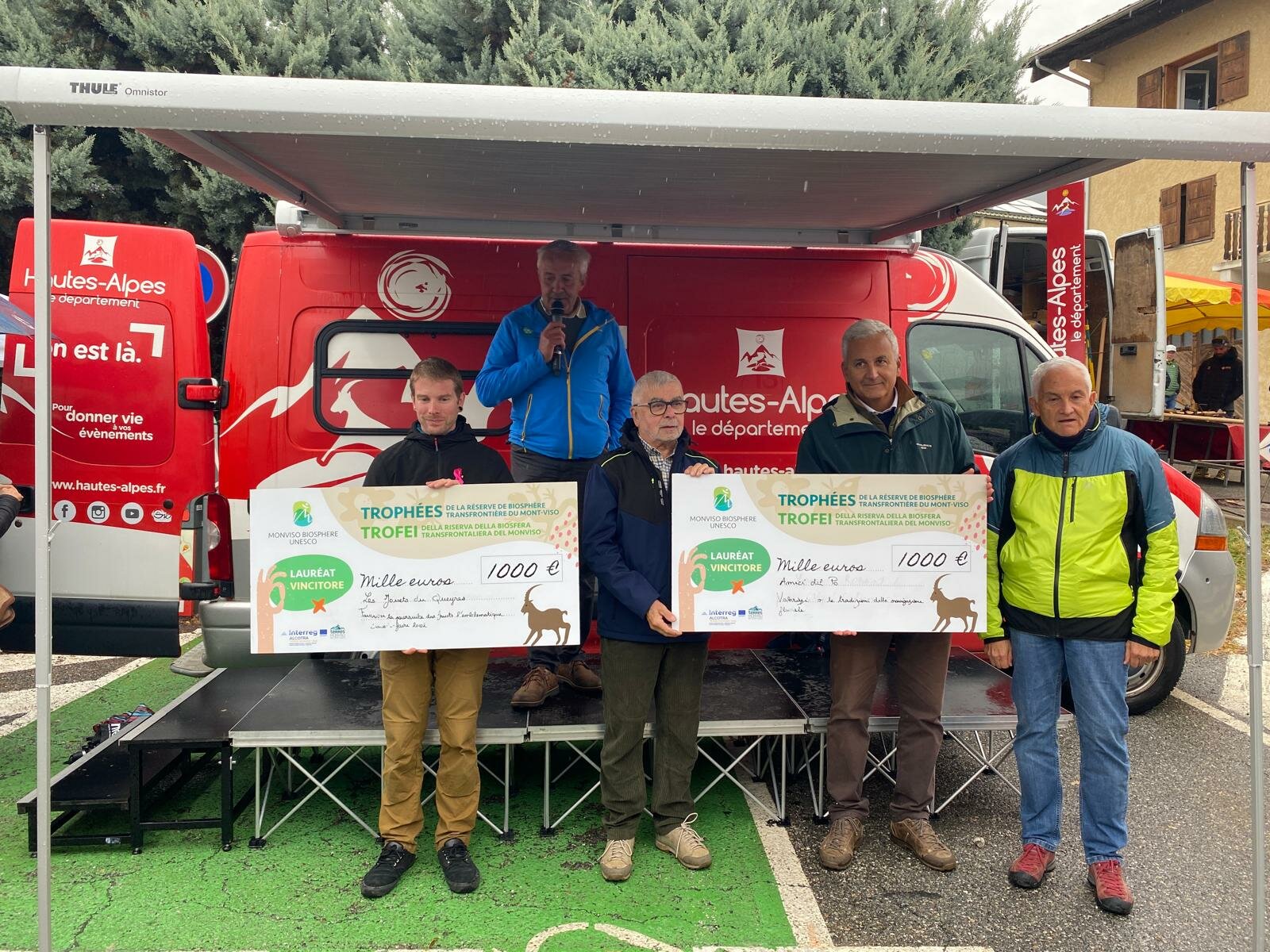 Nell’immagine sono ritratti cinque uomini su un piccolo palco all’aperto, davanti a un furgone rosso con il logo “Hautes-Alpes”. Due di loro tengono in mano due grandi assegni simbolici da 1.000 euro ciascuno, con l’intestazione “Trophées / Trofei della Riserva di Biosfera transfrontaliera del Monviso – Monviso Biosphere UNESCO”. Una persona al centro parla al microfono, durante la cerimonia di premiazione. L’atmosfera è conviviale e si riconosce un contesto di evento locale, con tende e altre persone sullo sfondo.