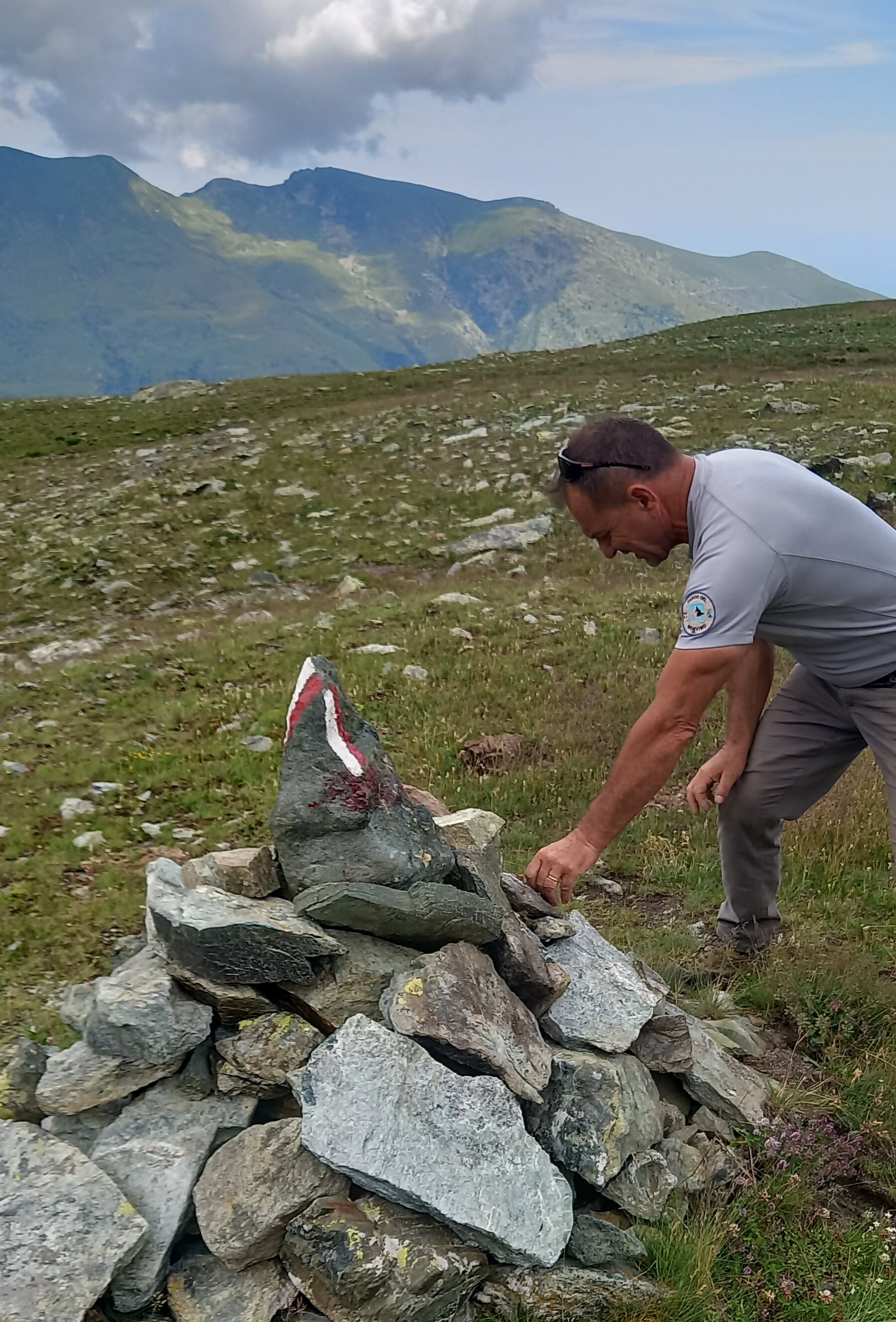 Un operatore del Parco del Monviso è inginocchiato accanto a un ometto di pietre lungo un sentiero di alta montagna, mentre sistema alcune rocce alla base della struttura. Sulla pietra sommitale è dipinto un segnavia bianco e rosso. Il paesaggio circostante è caratterizzato da pascoli d'alta quota, rocce sparse e montagne sullo sfondo, illuminate a tratti dalla luce che filtra tra le nuvole. L'immagine documenta un intervento di manutenzione della segnaletica escursionistica.