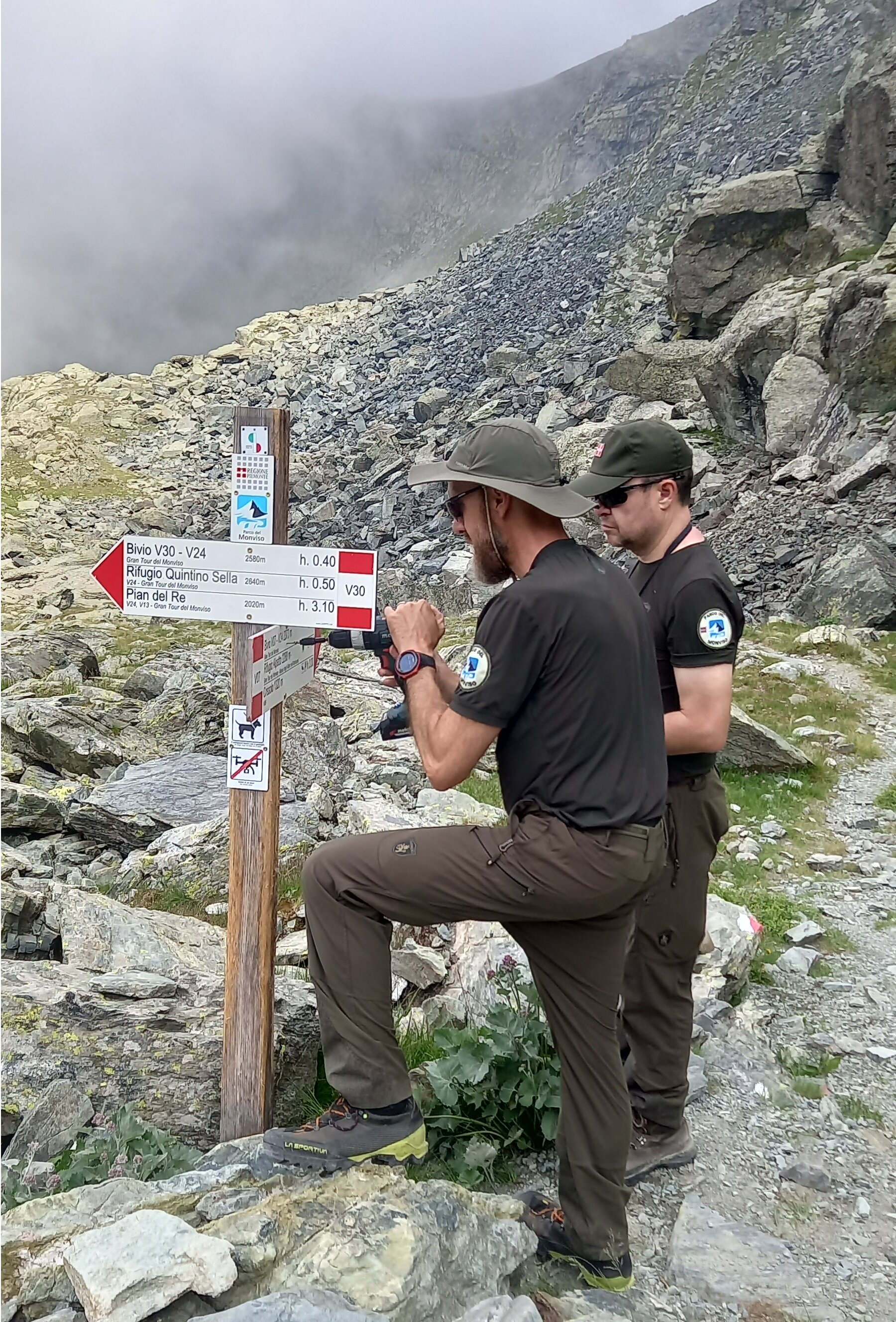 Due operatori del Parco del Monviso eseguono la manutenzione di un palo della segnaletica escursionistica in ambiente di alta montagna. Uno dei due utilizza un avvitatore per fissare una freccia direzionale, mentre il collega osserva il lavoro. I cartelli indicano il Rifugio Quintino Sella, il Pian del Re e il bivio per il Gran Tour del Monviso. Sullo sfondo si estende un paesaggio di rocce e detriti ai piedi di una parete montuosa, con nubi basse che avvolgono parzialmente le cime.