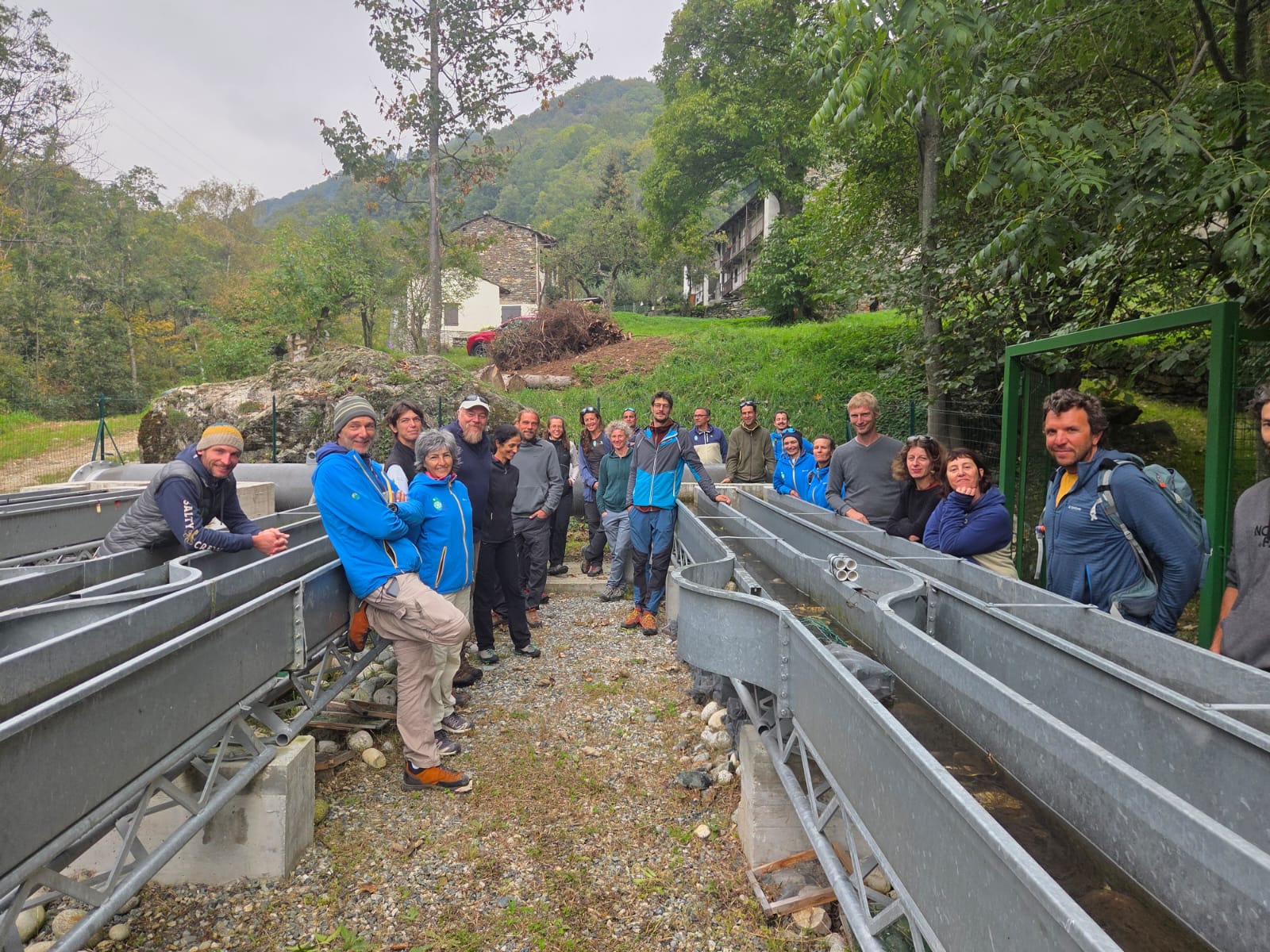 Gruppo di persone posa all’aperto tra lunghe vasche metalliche attraversate dall’acqua, in un impianto immerso nel verde. Sullo sfondo si vedono alberi, prati e alcune case in pietra di una borgata montana. Le persone indossano abbigliamento sportivo e da escursione e guardano verso la fotocamera.