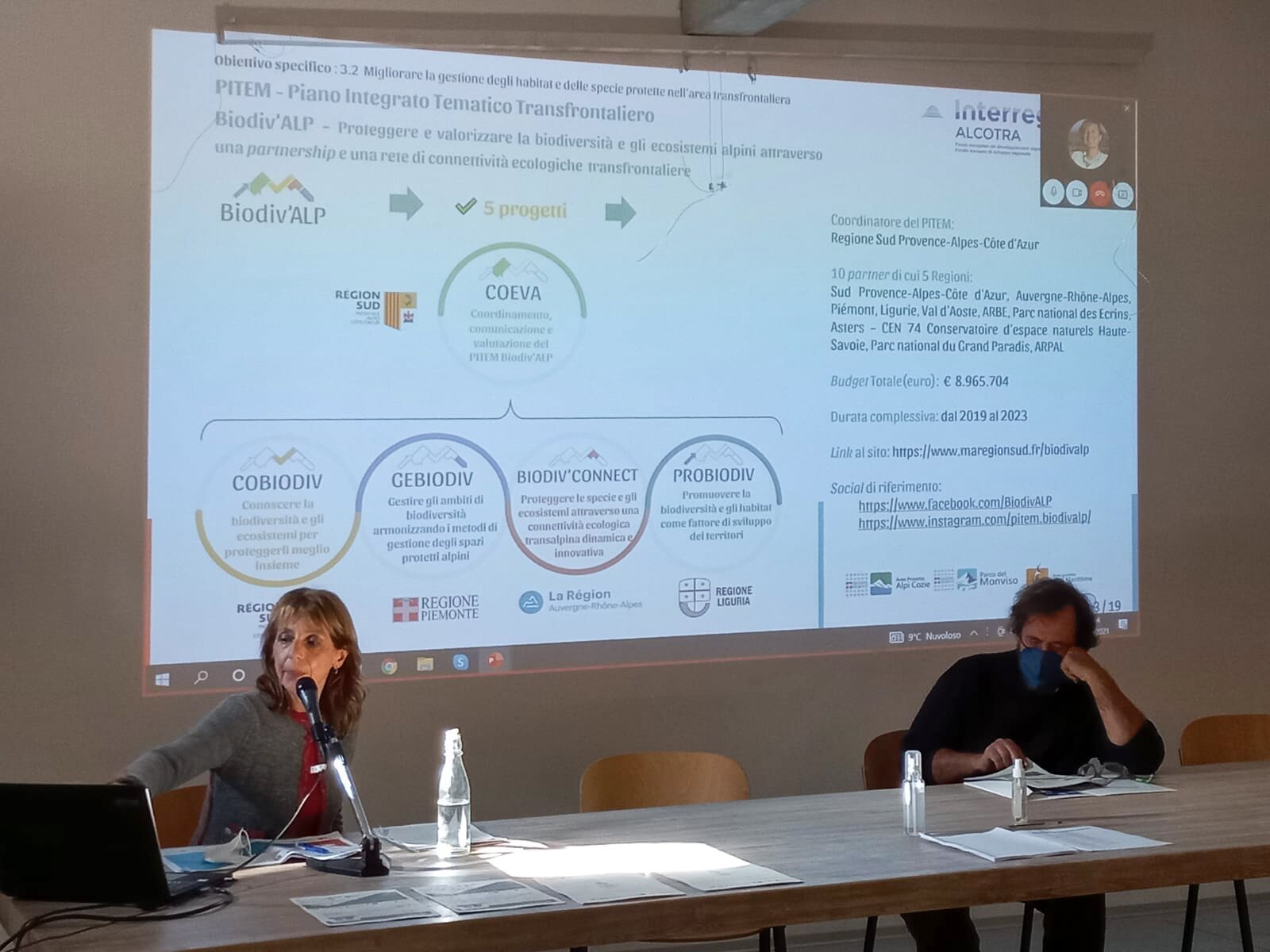 al banco dei relatori una donna e un uomo mentre alle loro spalle avanza la presentazione delle diapositive dell'incontro ed è in corso la connessione con un relatore collegato in videoconferenza remota