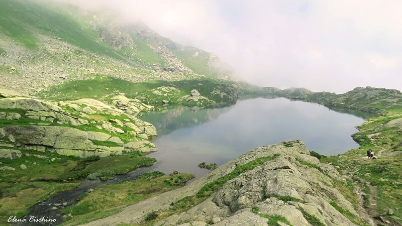 Lago alpino tra pendii erbosi e rocce, nuvole sullo sfondo
