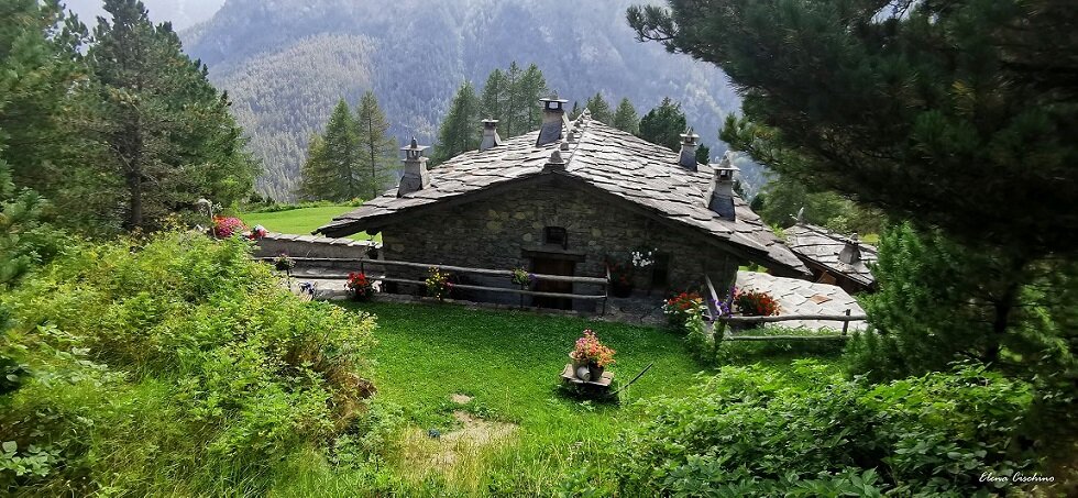 costruzione in pietra con tetto in lose con 5 camini e staccionata di legno con fiori sul davanti. un prato e alberi attorniano la costruzione