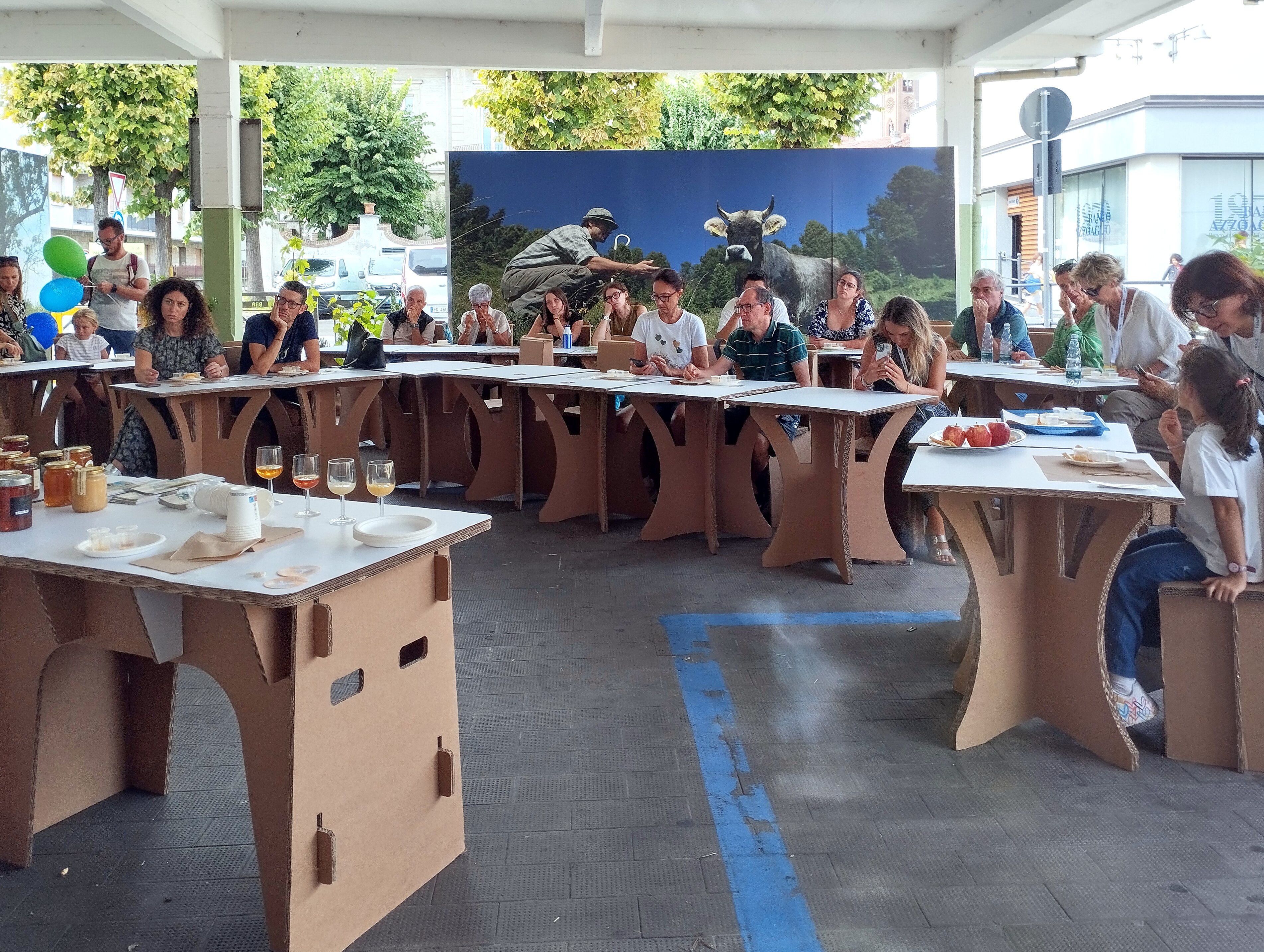 Una sessione di degustazione o workshop si svolge in uno spazio coperto con struttura a pilastri bianchi. Circa venti partecipanti di diverse età sono seduti lungo tavoli modulari in cartone ondulato color naturale, disposti a ferro di cavallo. I partecipanti, che includono uomini e donne di varie fasce d'età, stanno ascoltando attentamente e prendendo appunti. Sui tavoli sono visibili bicchieri con liquidi di colore ambrato (probabilmente miele o idromele), piatti bianchi, vasetti di miele e materiale informativo. Sullo sfondo domina un grande pannello fotografico che mostra un pastore in abiti tradizionali accanto a una mucca pezzata in un paesaggio montano verdeggiante. L'ambiente è luminoso grazie alle ampie aperture che si affacciano su una strada alberata con edifici residenziali. Sul pavimento scuro sono visibili alcune strisce blu di demarcazione. L'atmosfera è quella di un evento formativo legato ai prodotti locali e all'agricoltura di montagna.