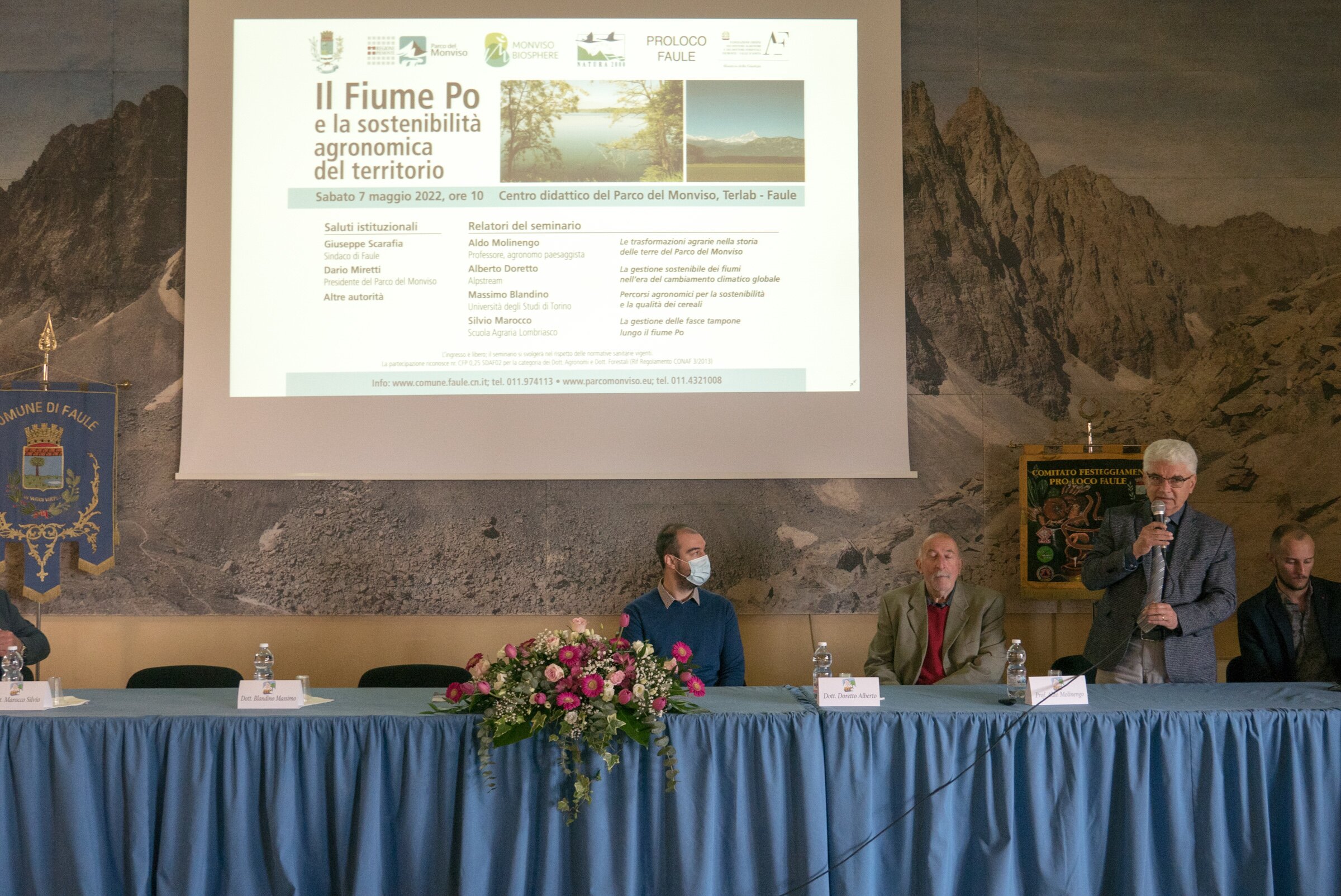 Tre persone dietro a un tavolo relatori, al di sopra viene proiettata la locandina del seminario in corso