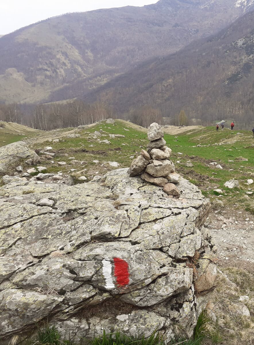 Panorama verso la bassa valle, in primo piano una roccia con segnaletica sentieri in vernice, in secondo piano tre escursionisti che scendono lungo un sentiero