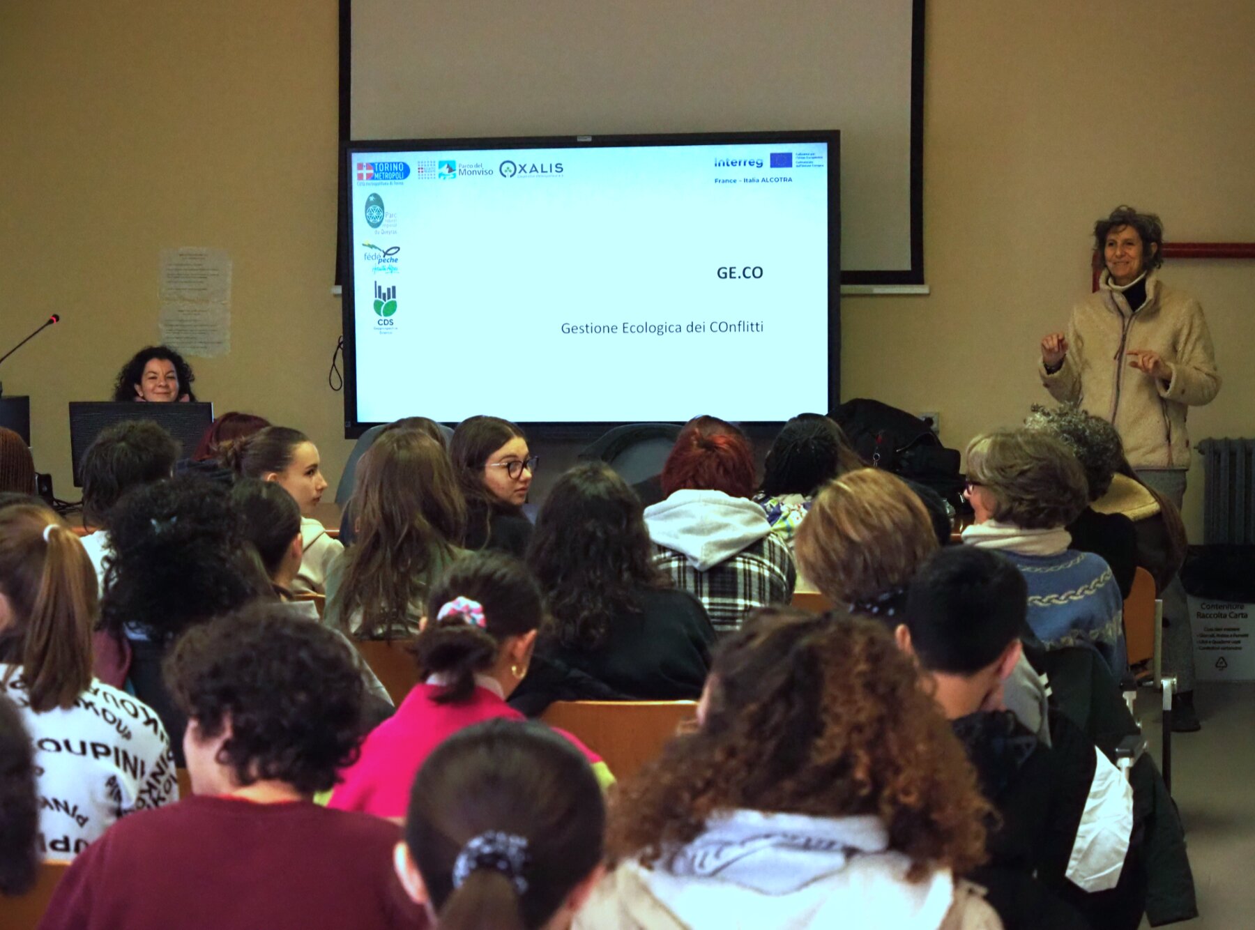 Una docente sta parlando ad una classe di studenti, posti di spalle rispetto al punto in cui è stata scattata la foto.