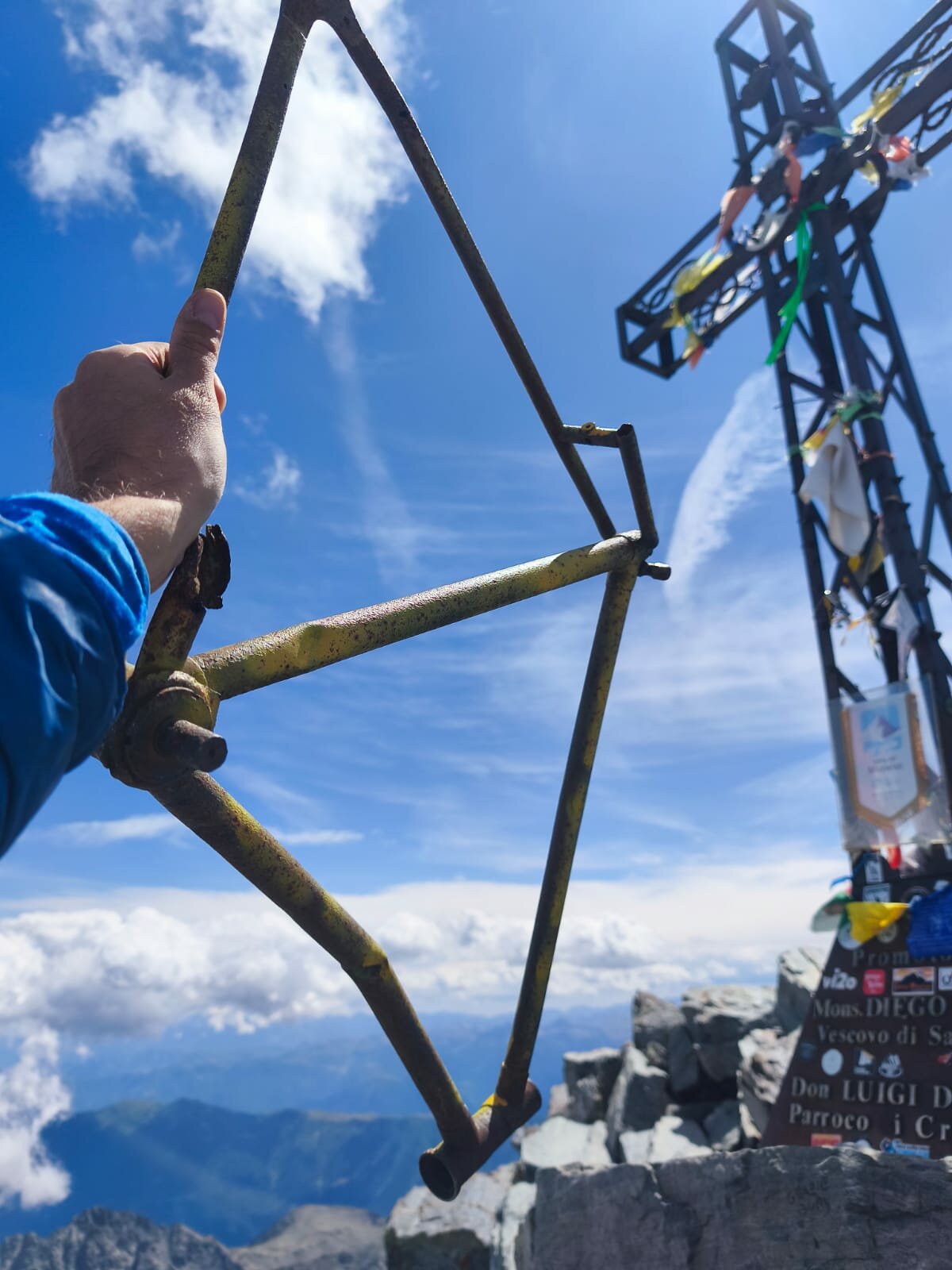 a sinistra si vede una mano che sorregge il telaio della bicicletta, sulla destra la croce di vetta del Monviso. Sullo sfondo nuvole e le montagne sottostanti.