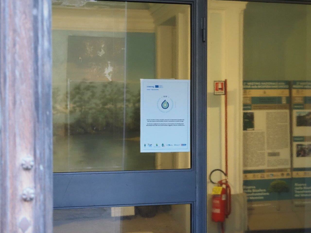 una porta a vetri, l'ingresso dell'Ente di Gestione delle Aree protette del Monviso, sulla quale è stato affisso il manifesto del progetto GE.CO