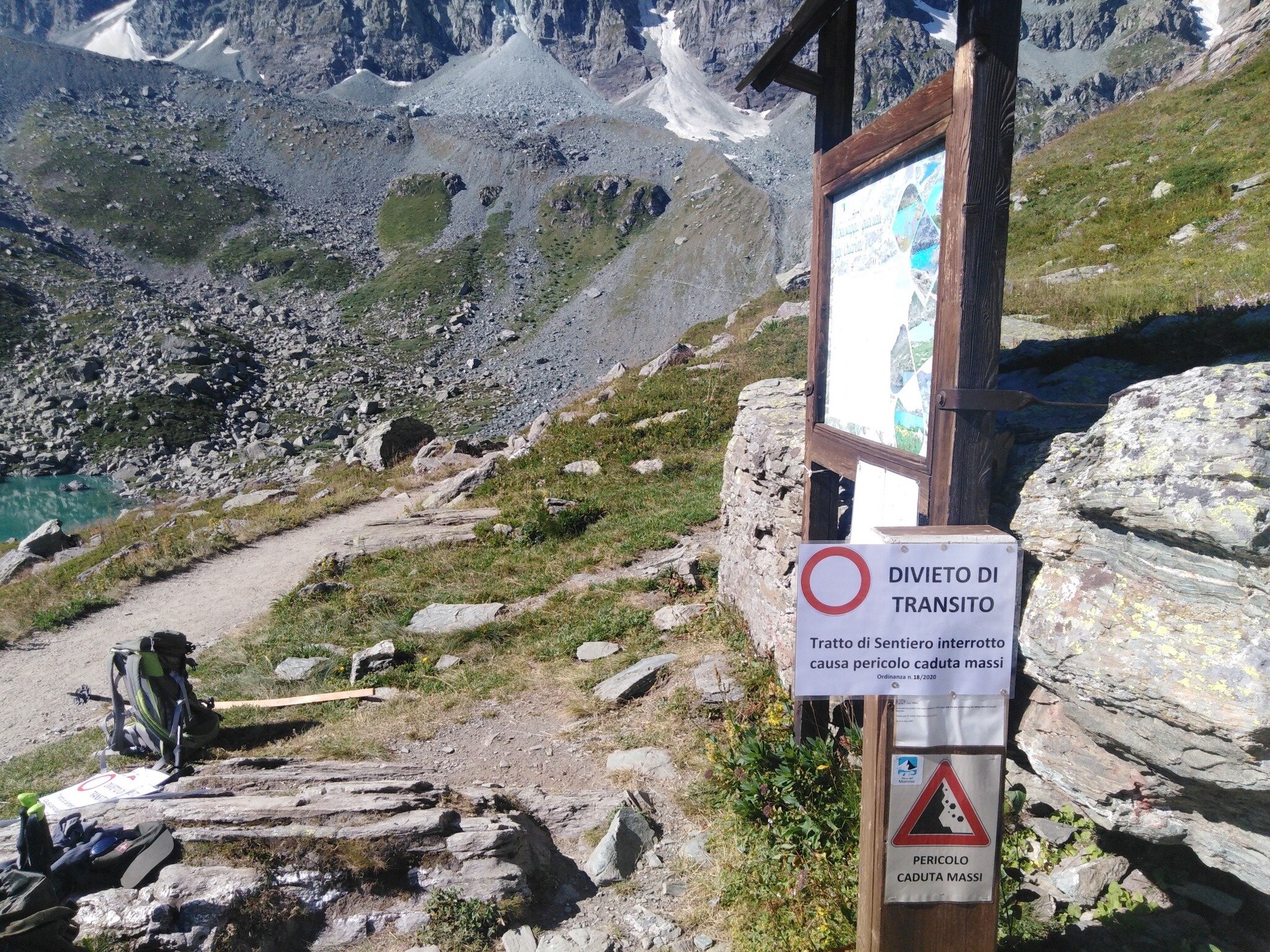 sulla sinistra della foto, uno zaino e uno scorcio del lago Chiaretto, sulla destra il cartello di divieto di transito attaccato alla bacheca con la mappa del sentiero CNAV13.