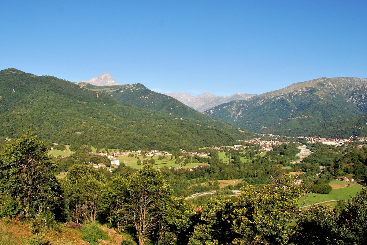 Panorama su una valle verdeggiante, con un abitato in lontananza dietro al quale si rialzano i rilievi; in secondo piano la punta del Monviso.