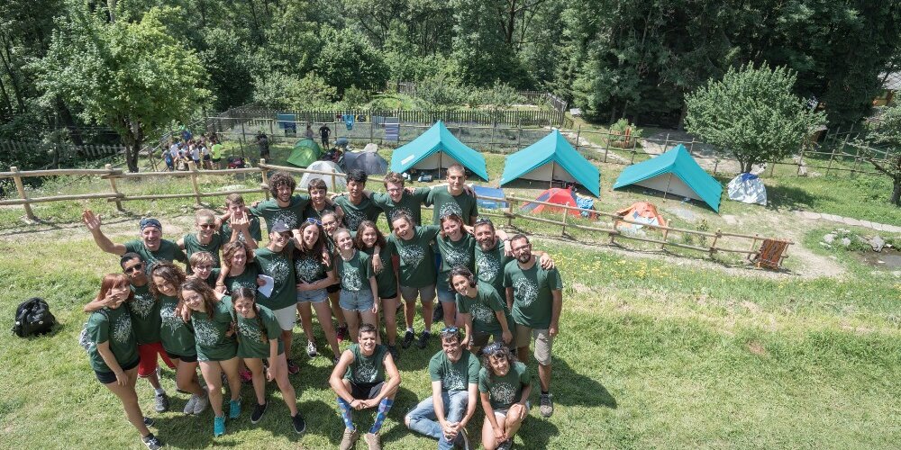 Foto di gruppo di 30 ragazzi con maglietta verde del MaB Camp. Sullo sfondo tre tende da campeggio in un prato con alberi