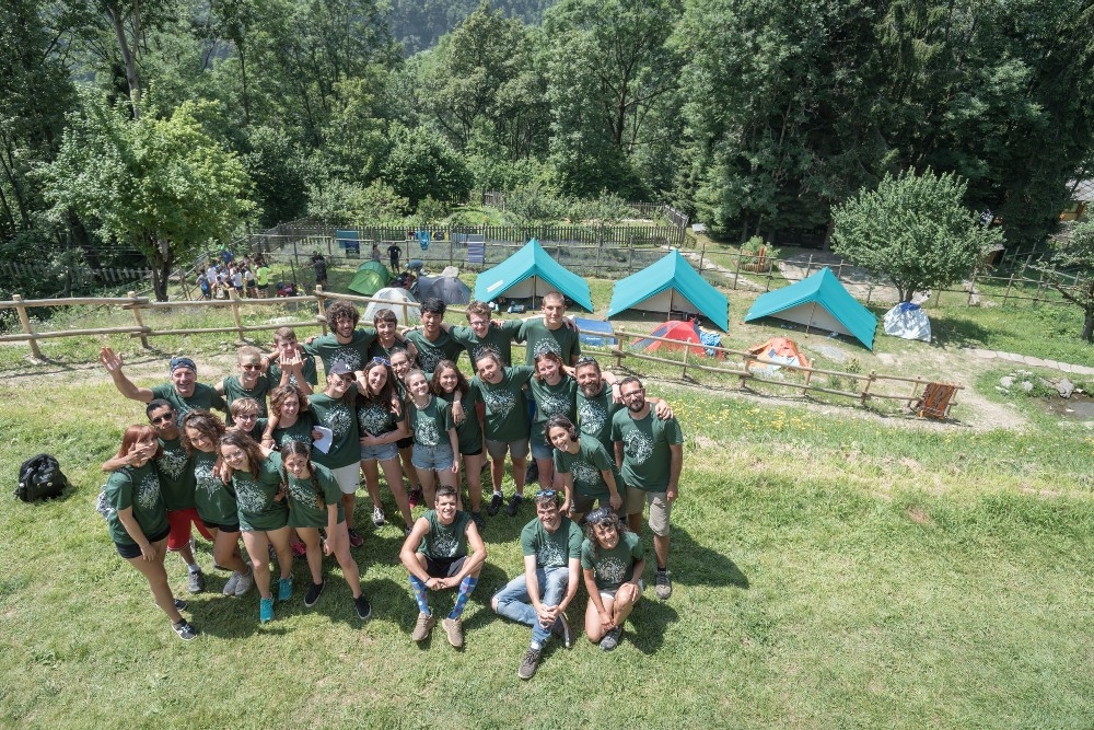 Foto di gruppo di 30 ragazzi con maglietta verde del MaB Camp. Sullo sfondo tre tende da campeggio in un prato con alberi
