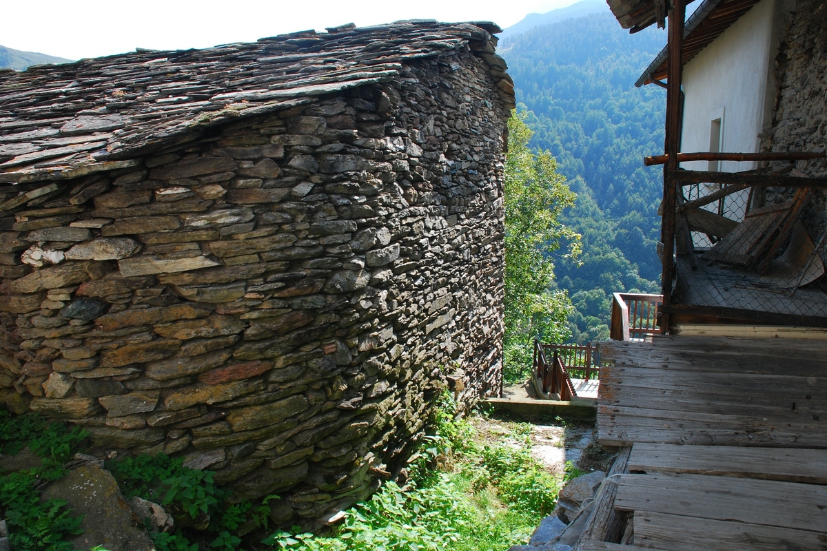 in primo piano una casa di pietra con tetto a lose, sulla destra un balcone in legno. Sullo sfondo montagne con boschi
