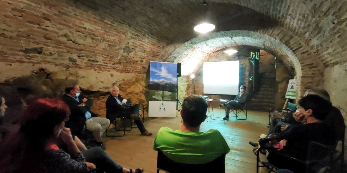 Gruppo di persone sedute in cerchio in un locale con volta a mattoni del Castello della Manta, presente un roll-up con l'immagine della Riserva della Biosfera transfrontaliera del Monviso