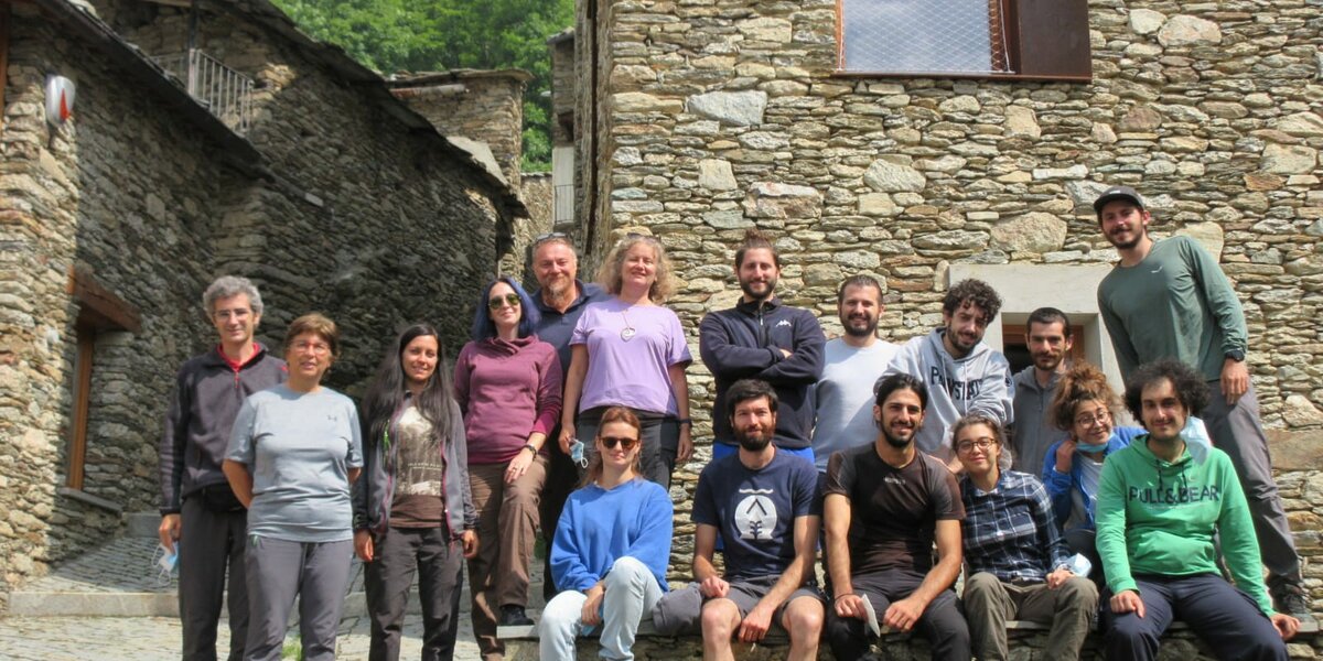 Diciassette persone, uomini e donne su due file, una in piedi e l'altra seduta, in un borgo montano con muri a secco