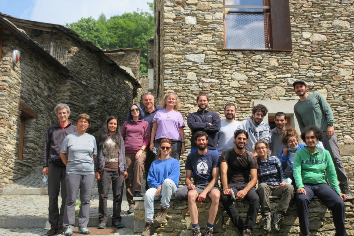Diciassette persone, uomini e donne su due file, una in piedi e l'altra seduta, in un borgo montano con muri a secco