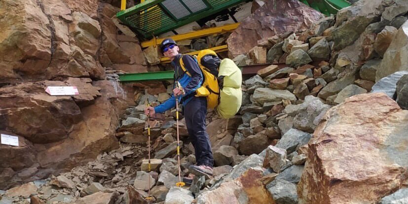 bivacco alpino in lamiera su parete ripida che sale verso il Monviso. Un alpinista in primo piano con due zaini