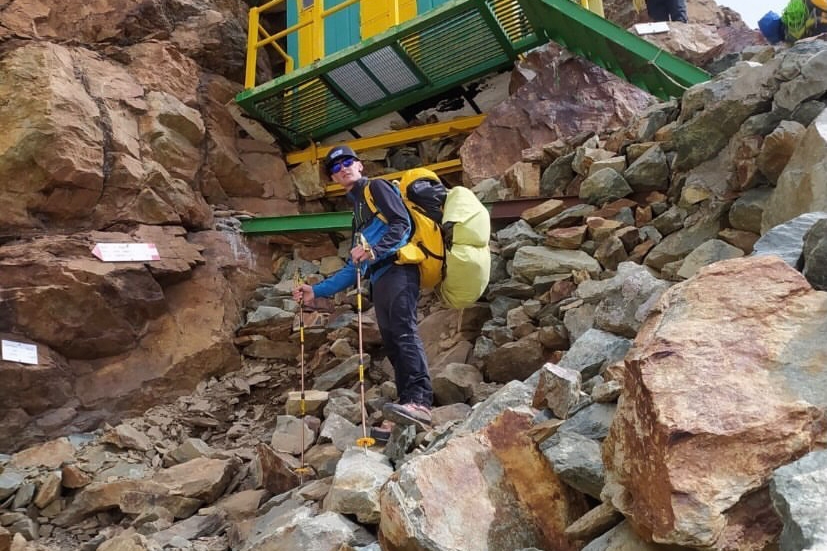 bivacco alpino in lamiera su parete ripida che sale verso il Monviso. Un alpinista in primo piano con due zaini