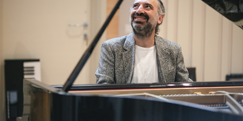 Stefano Bollani sorridente seduto al pianoforte a coda con lo sguardo rivolto verso l'alto