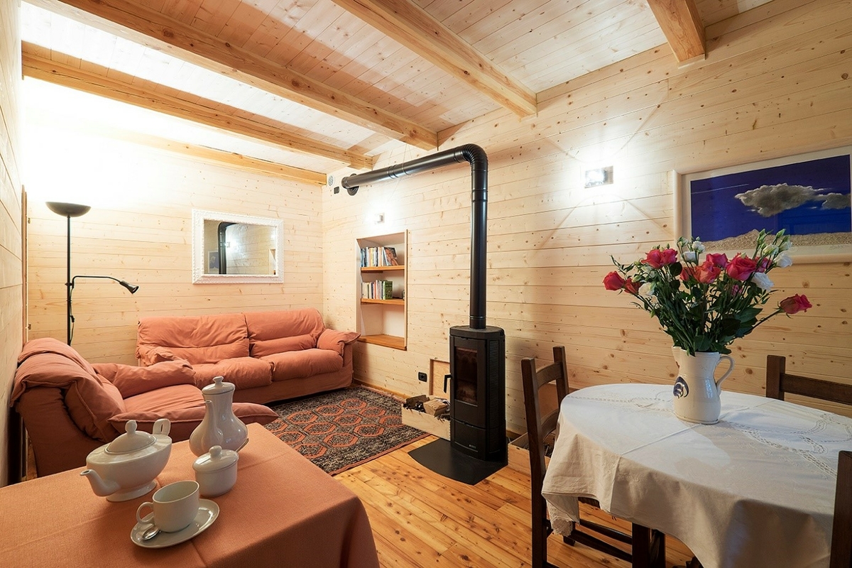 Interno di spazio comune del B&B con pareti, pavimenti e soffitti in legno. in primo piano a sinistra una tavolo con tovaglia rosata con servizio da the bianco, a destra tavolo rotondo con tovaglia bianca, tre sedie e vaso con rose. in secondo piano stufa a legna e salottino con due divani, tappeto e libreria