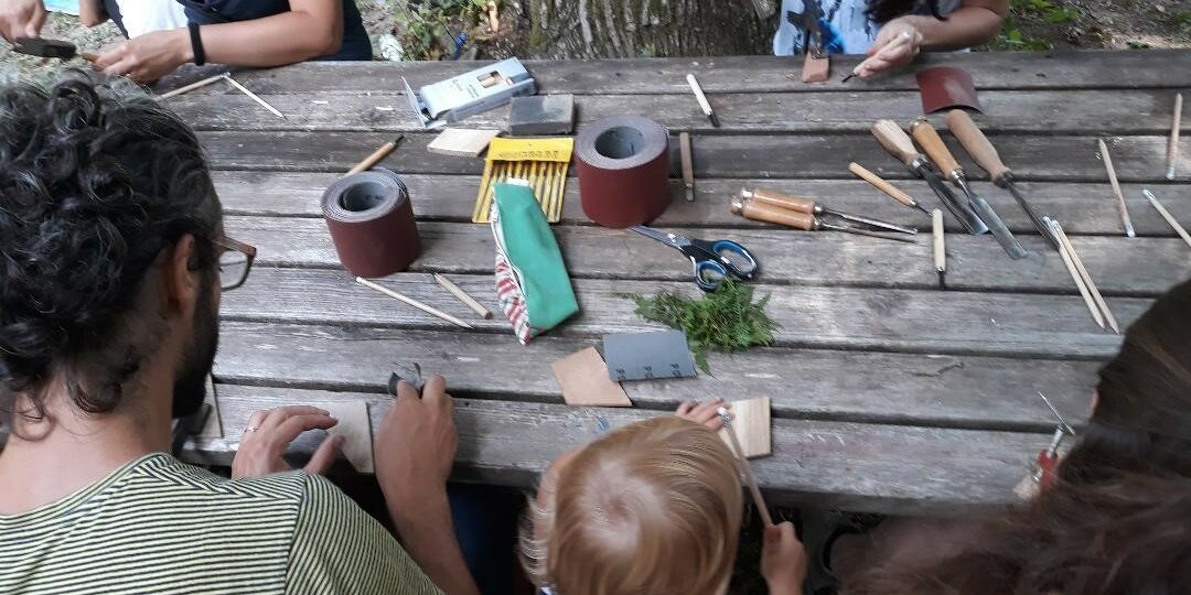 adulti e bambini si cimentano in un laboratorio sul legno. seduti a un tavolo di legno all'esterno; sul tavolo attrezzi e pezzi di legno.