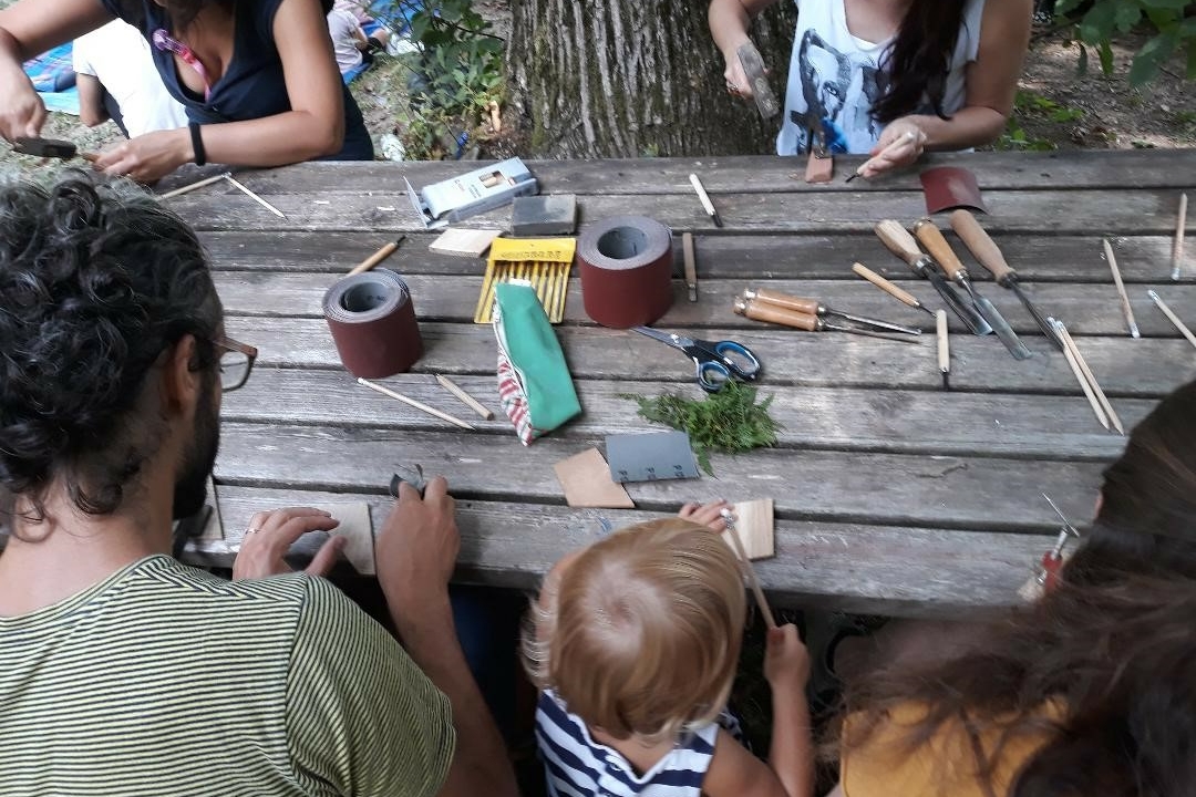 adulti e bambini si cimentano in un laboratorio sul legno. seduti a un tavolo di legno all'esterno; sul tavolo attrezzi e pezzi di legno.