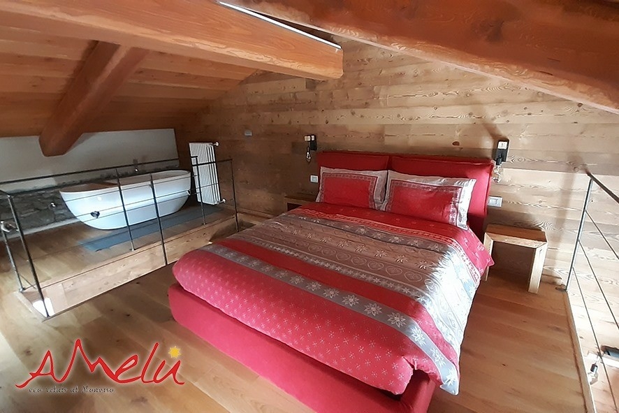 In una mansarda con tetto e pareti in legno un letto matrimoniale con piumone rosso e una vasca da bagno sulla sinistra