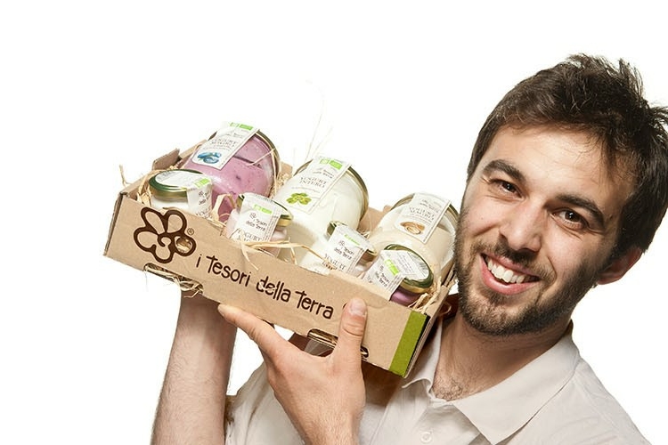 Un ragazzo sorridente porta in spalla una cassetta piena di vasetti con la scritta I tesori della Terra